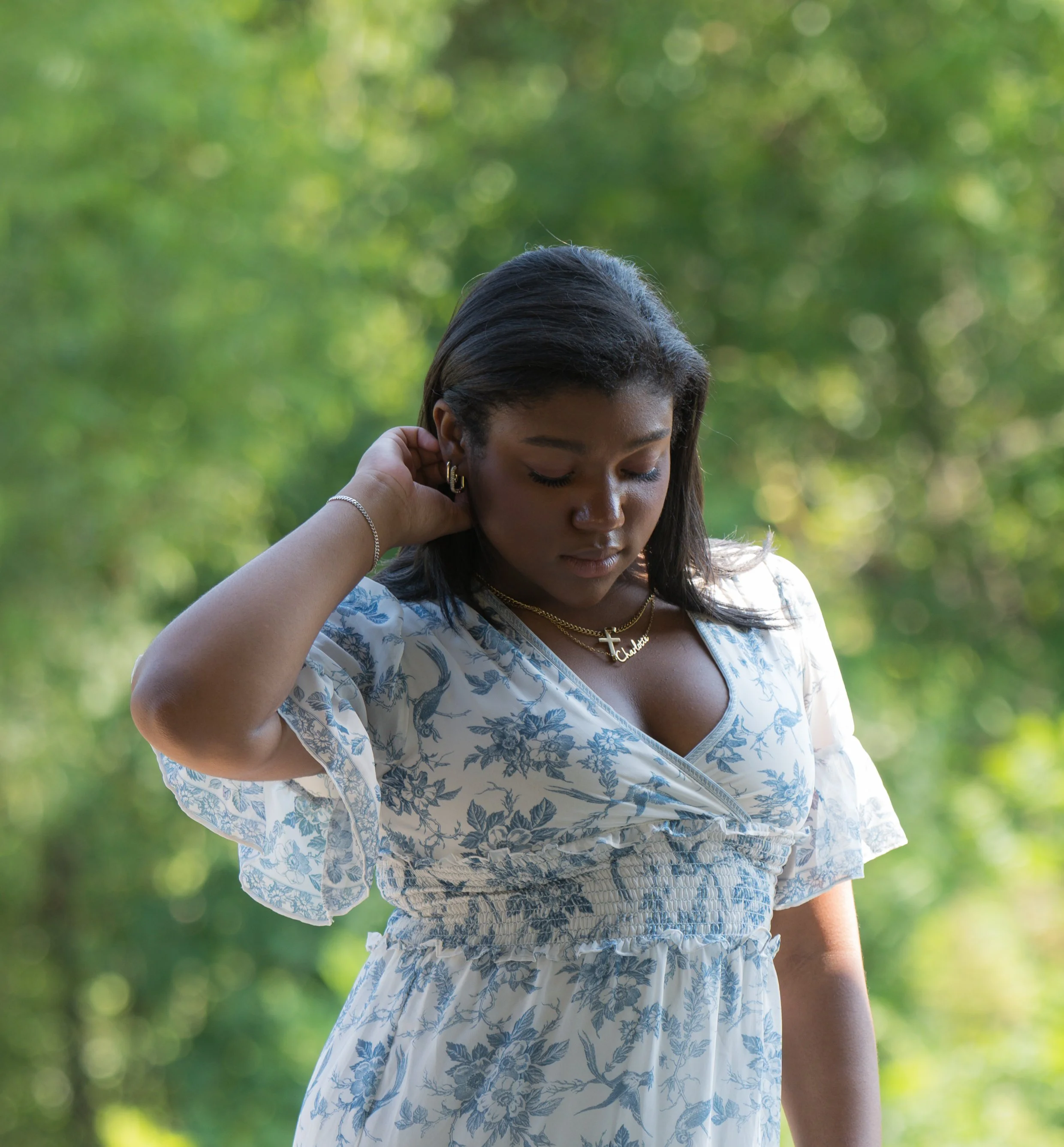 A young woman with dark hair wearing a white dress with blue floral patterns, standing outdoors with a blurred green background. She is looking down, touching her hair with her right hand, and wearing jewelry including a cross necklace, earrings, and