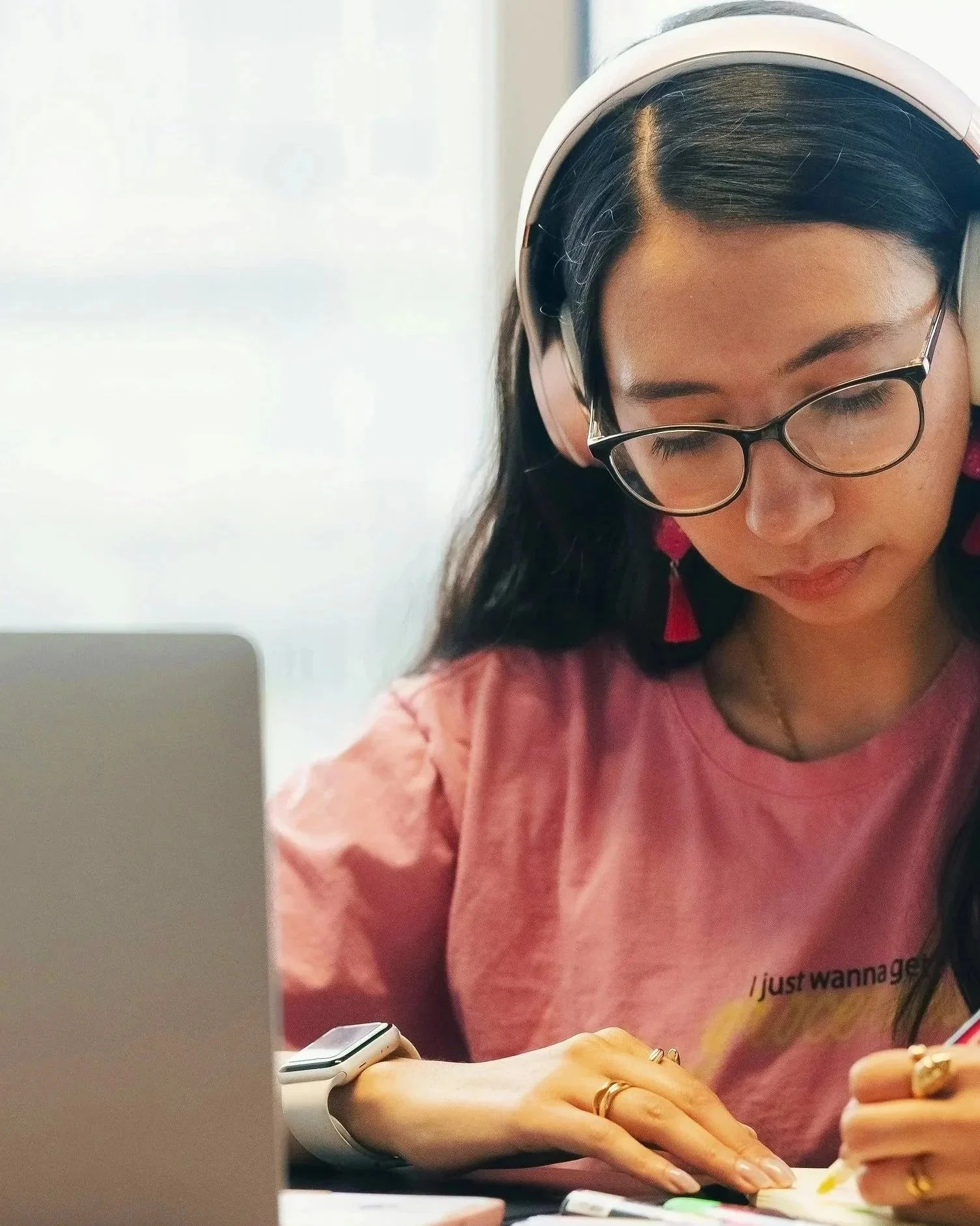 A young woman with glasses and long dark hair, wearing pink earrings, a pink T-shirt, and an Apple Watch, is working at a desk in front of a computer.
