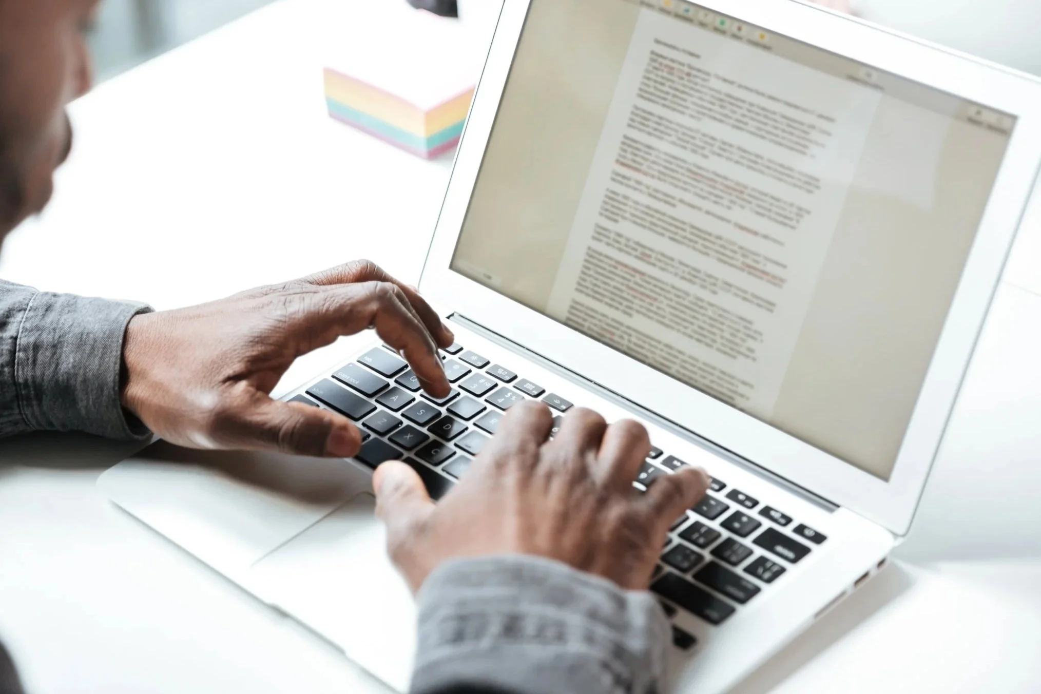 Person typing on a silver laptop with a document on the screen, beside a colorful sticky note pad.