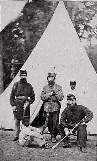 Four men dressed in historical military uniforms posing in front of a teepee tent, with a dog lying on the ground.