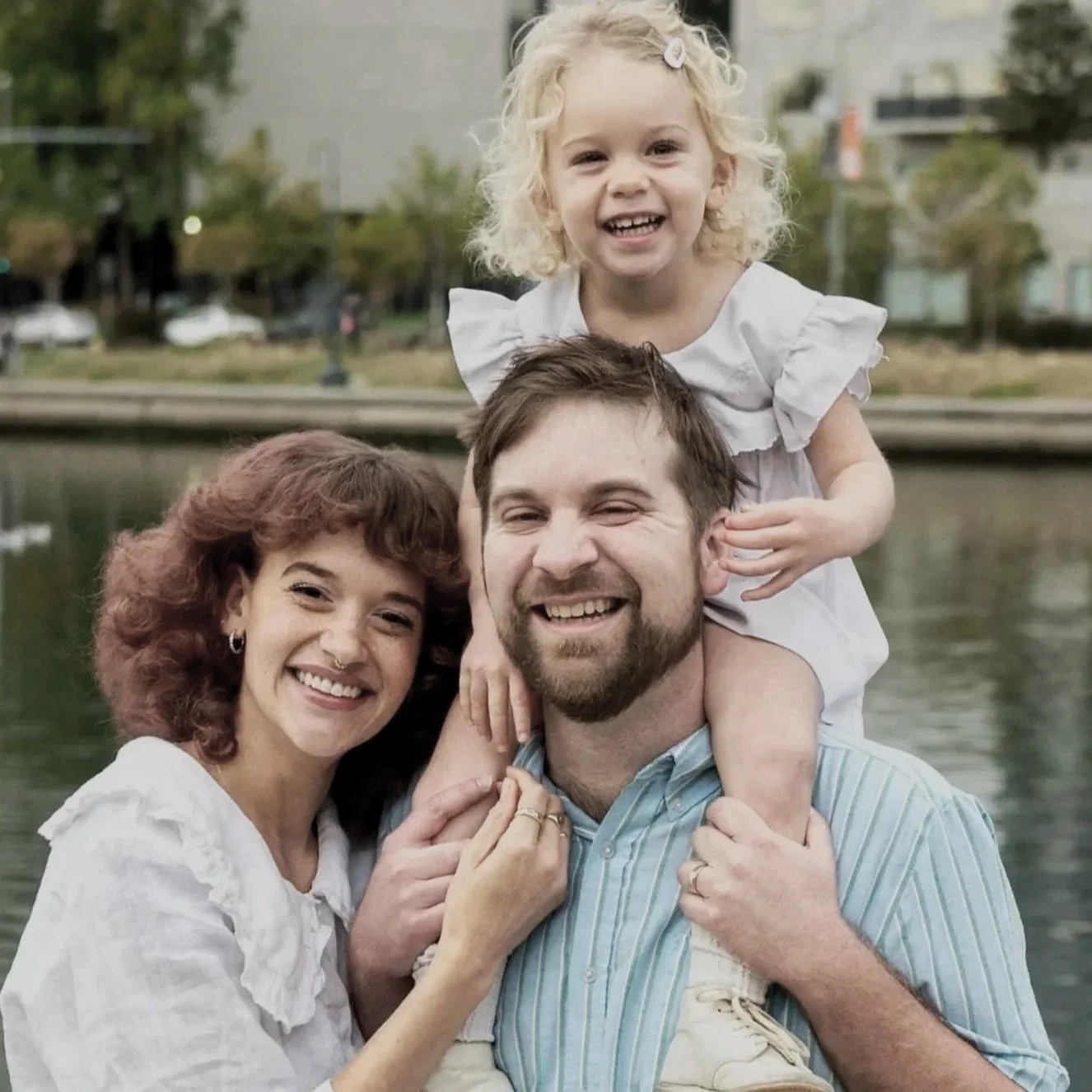 A happy family of three smiling by a body of water during daytime. The family includes a woman with curly red hair, a man with short brown hair and a beard, and a young girl with blonde curly hair sitting on the man's shoulders.