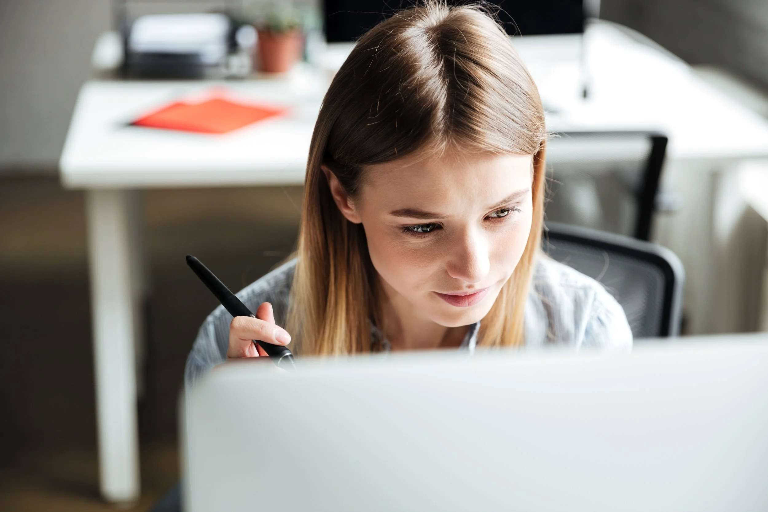 A young woman with brown hair working at a computer with a pen in her hand, in a well-lit office.