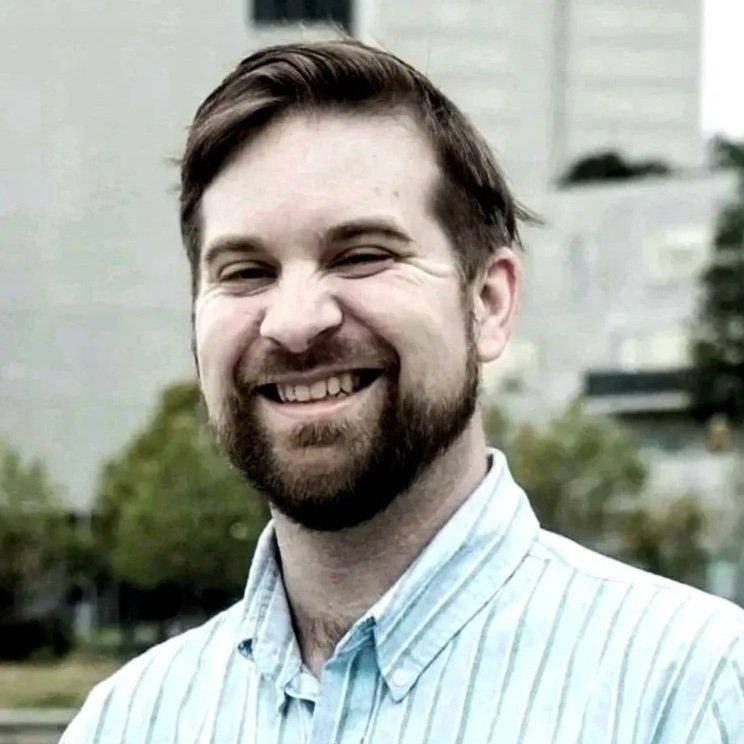 A smiling man with a beard wearing a light blue and white striped button-up shirt, standing outdoors with blurred trees and buildings in the background.