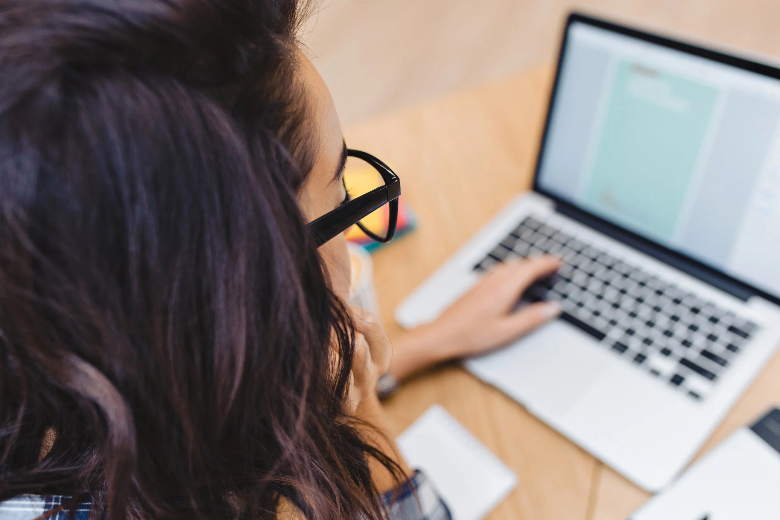 A woman with glasses and long dark hair working on a laptop at a wooden desk.