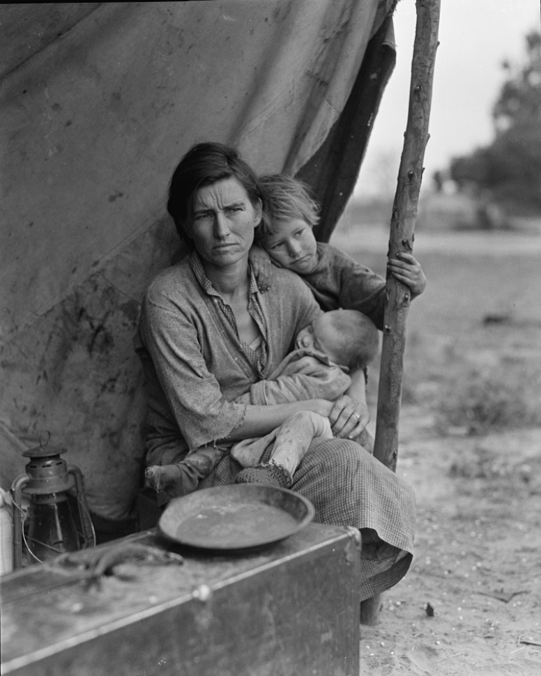 A woman sitting outside a tent holding a baby, with a young boy leaning on her shoulder, in a rural setting.