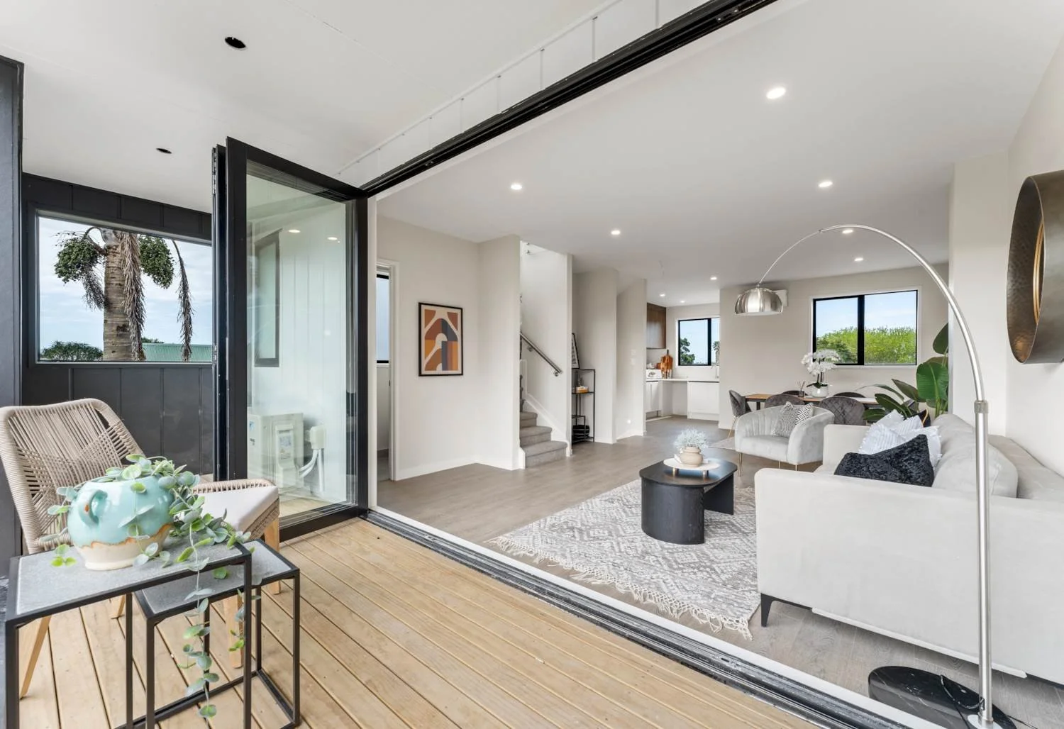 A modern living room and dining area featuring light-coloured flooring and large glass bi-fold doors that open onto a wooden balcony, creating a seamless indoor-outdoor flow. 