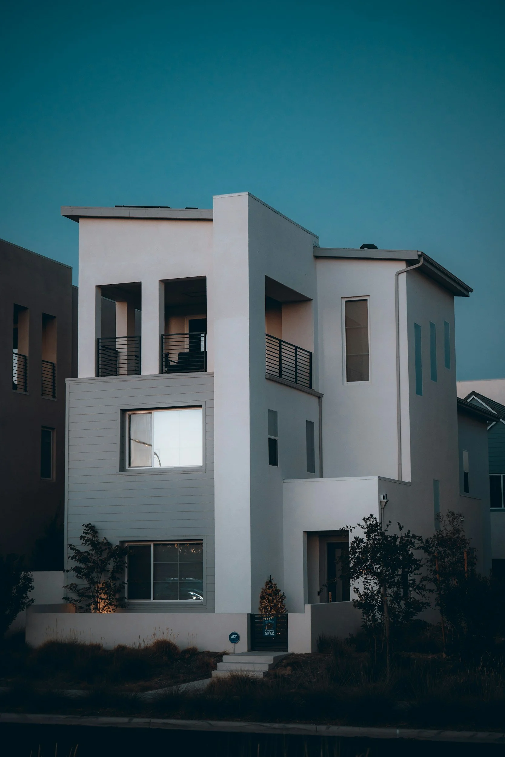 Exterior view of a modern multi-story residential home at dusk, featuring a contemporary white and grey facade, large windows, and a top-floor balcony.