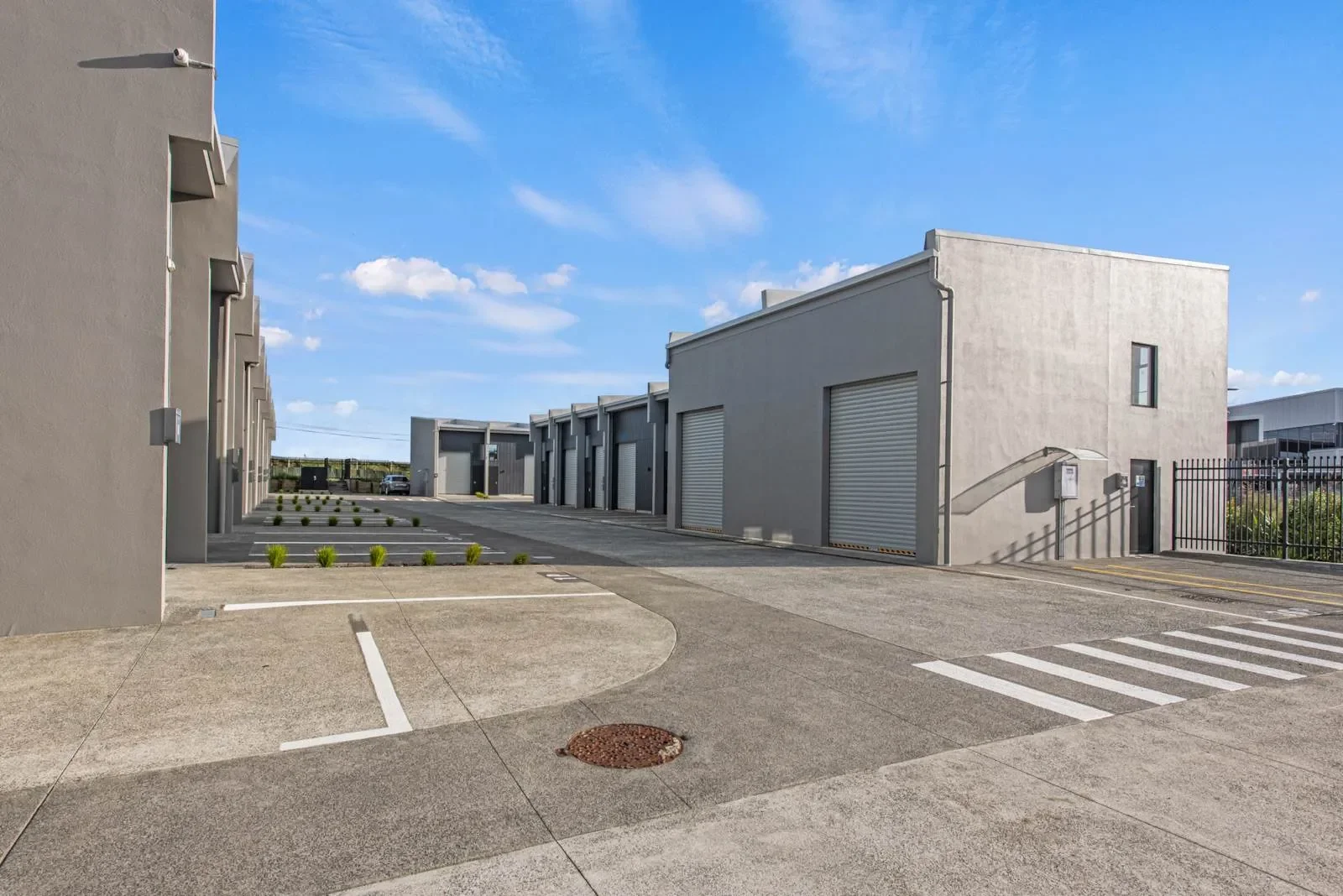 Exterior view of a modern industrial property featuring multiple grey units with secure roller doors. The business complex includes a paved concrete courtyard with clearly marked parking spaces and access routes.