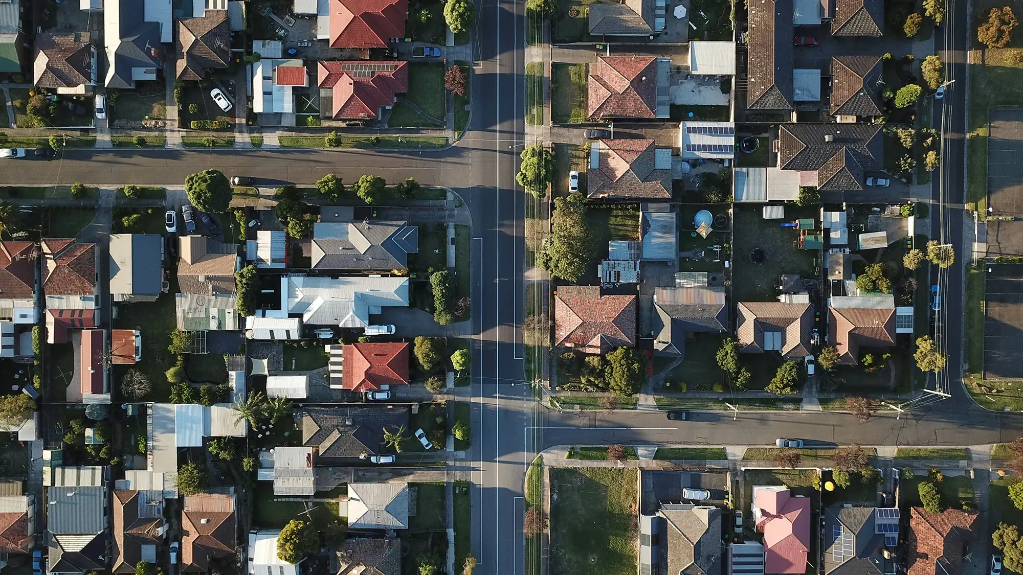 An aerial view of a residential neighborhood with houses, driveways, trees, and streets. Some houses have solar panels on the roofs, and there are parked cars along the streets and in driveways.