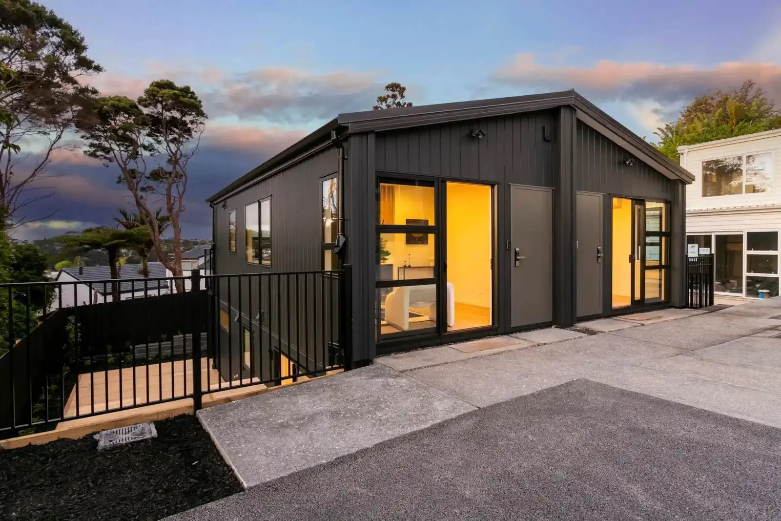 Exterior street view of modern dark grey townhouses featuring glowing glass doors, a concrete driveway, and black metal fencing at dusk.