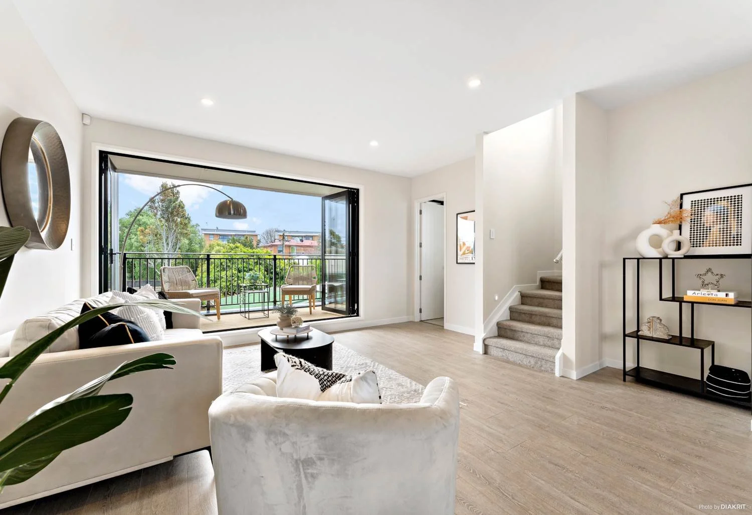 An open-plan living room interior featuring light wood floors, contemporary furnishings, and a carpeted staircase. Large bifold glass doors seamlessly connect the indoor space to the balcony, maximising natural light and indoor-outdoor flow.