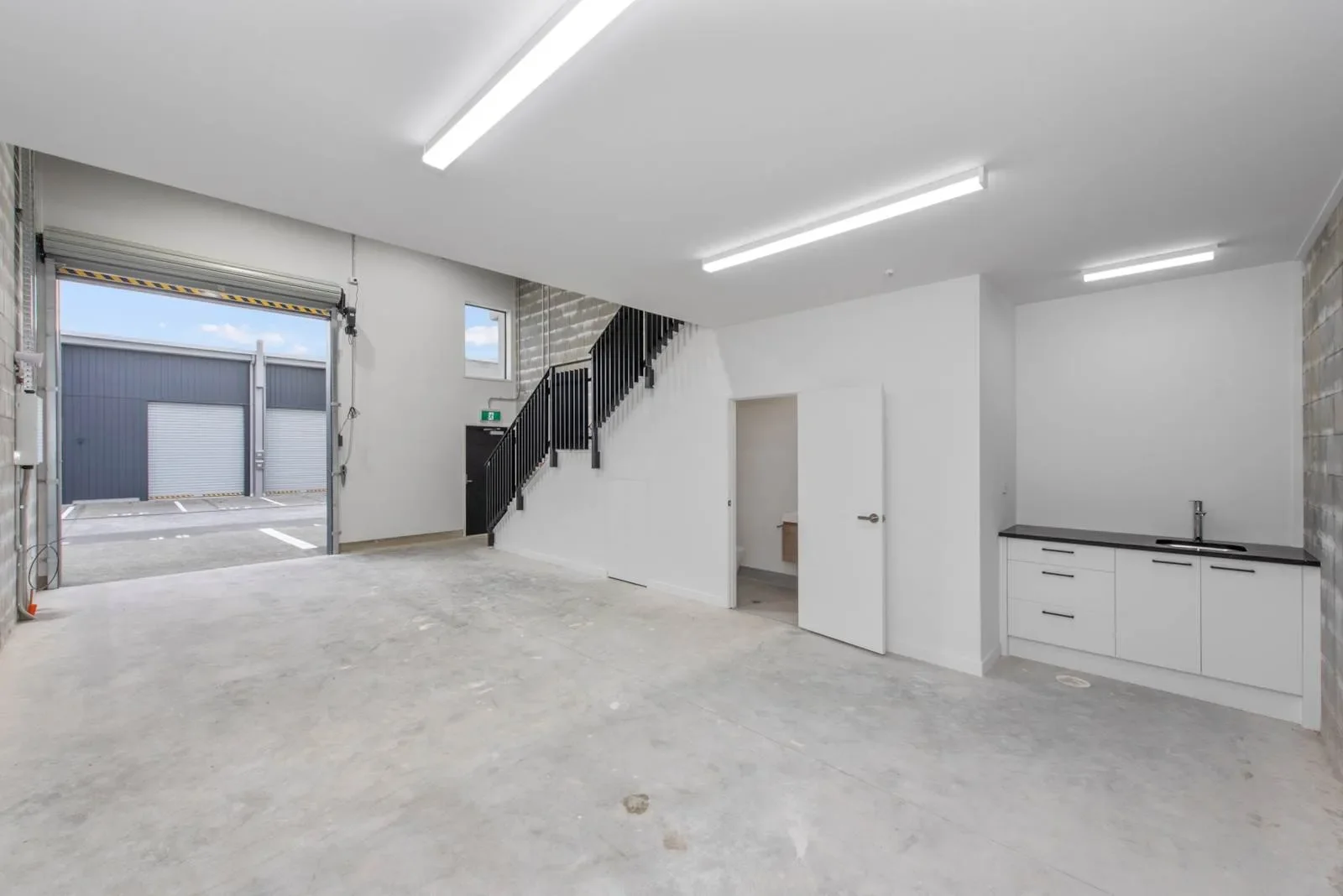 Interior view of a versatile modern industrial warehouse unit featuring a spacious concrete floor, a built-in kitchenette, and an open roller door. Also, it includes a black metal staircase and accessible bathroom facilities.
