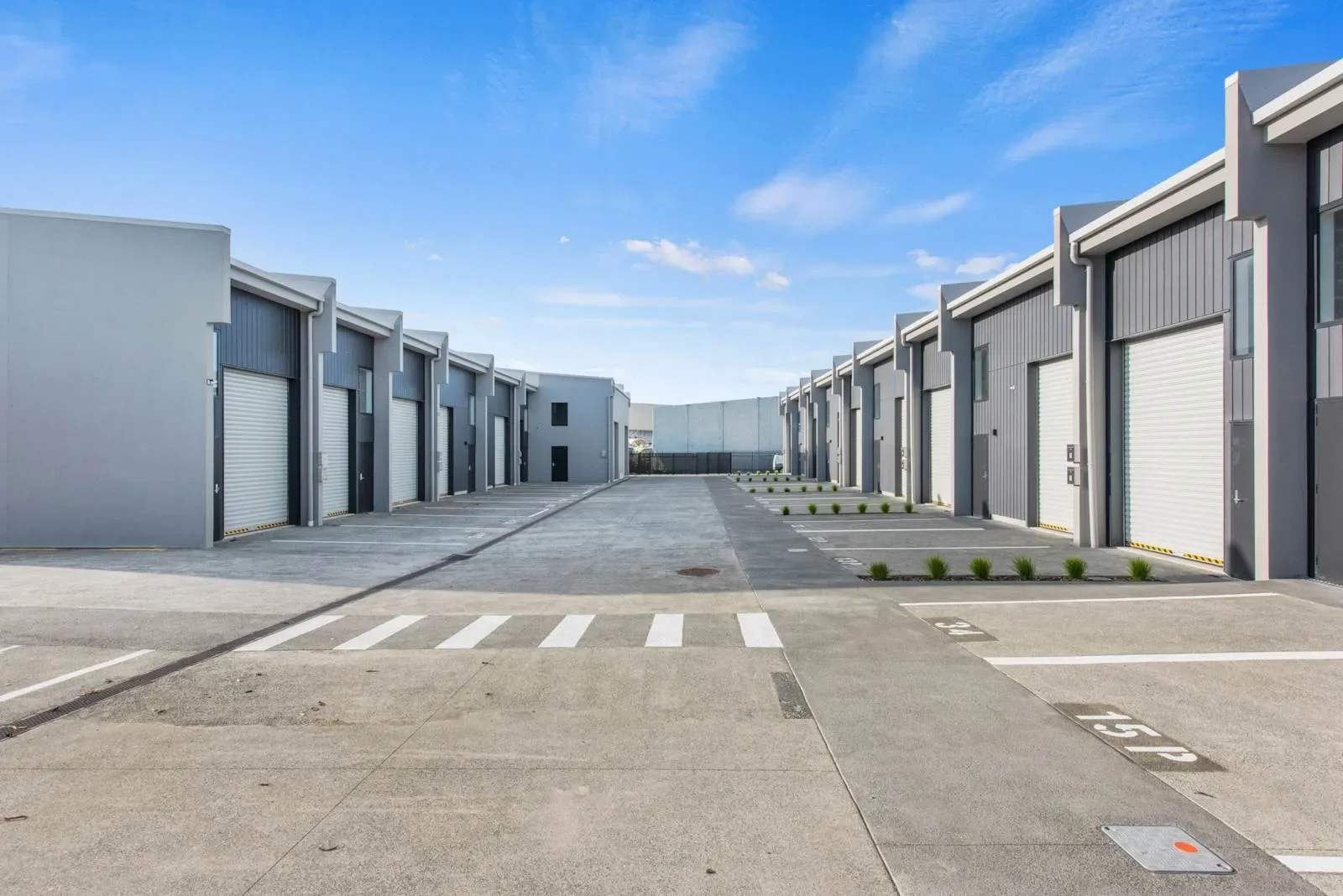 Exterior view of a modern industrial business park featuring parallel rows of grey commercial warehouse units with white roller doors. A wide concrete driveway with marked parking spaces provides clear access to the facilities.