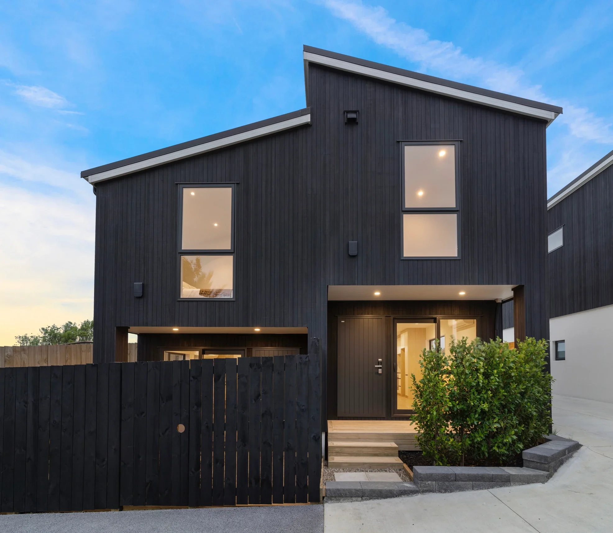 Exterior view of a modern two-story residential home in Torybay, Auckland, featuring dark vertical cladding and a slanted roof. The warmly illuminated entrance showcases a wooden porch and is enclosed by a contemporary black timber fence.