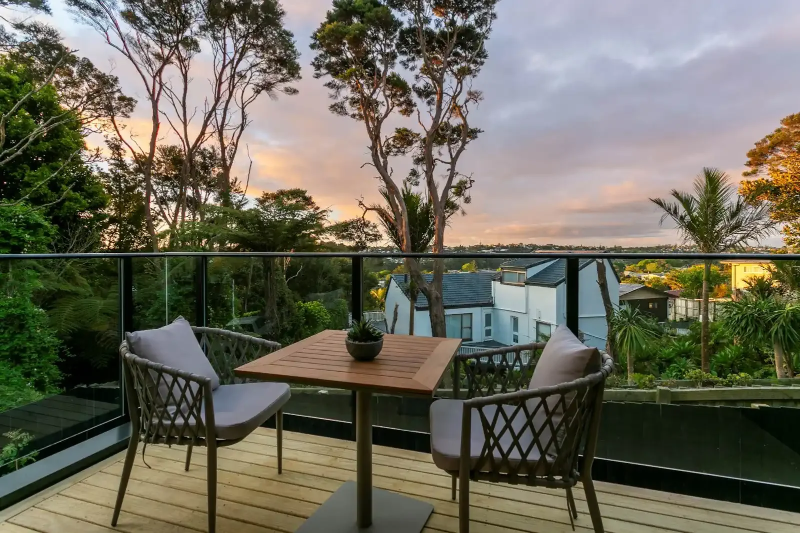 A modern outdoor balcony with a wooden deck and glass railing, featuring a small patio table and two woven chairs overlooking lush trees and residential homes during a scenic sunset.
