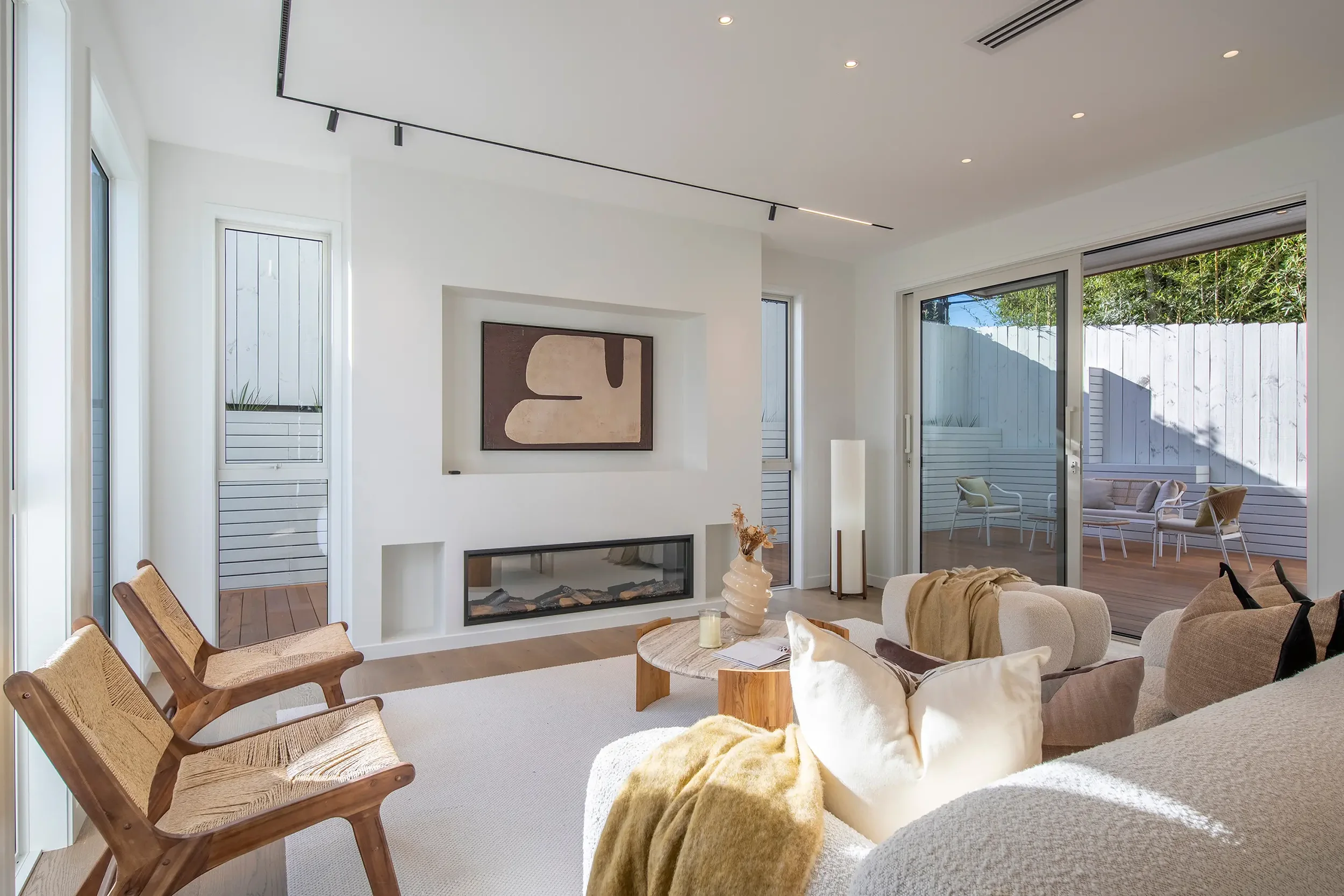 A spacious outdoor wooden deck attached to a modern dark grey house, featuring sliding glass door that open into an illuminated bedroom at dusk.