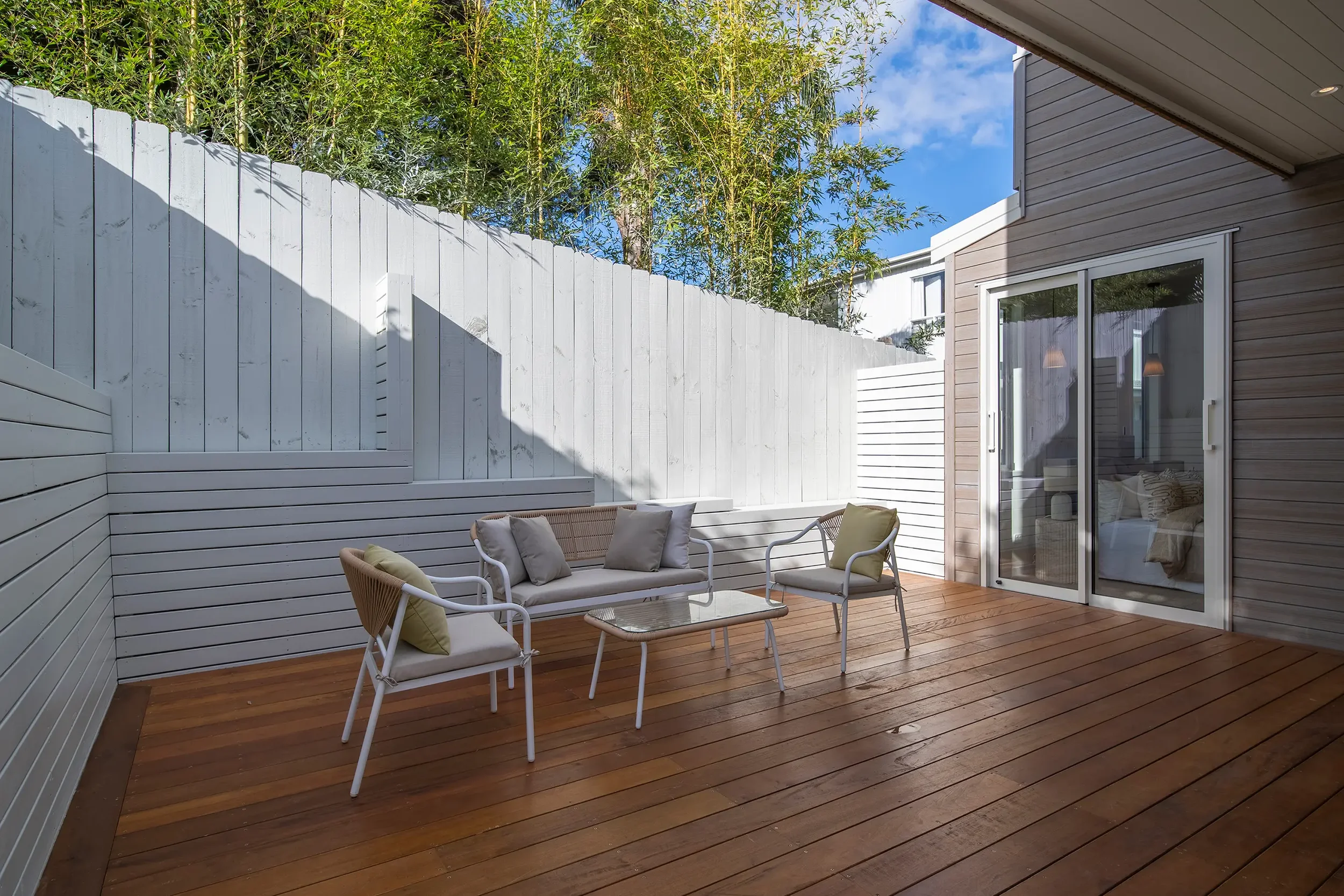 A modern outdoor balcony featuring a wooden deck, glass railings, and a small dining set overlooking lush green trees at sunset.