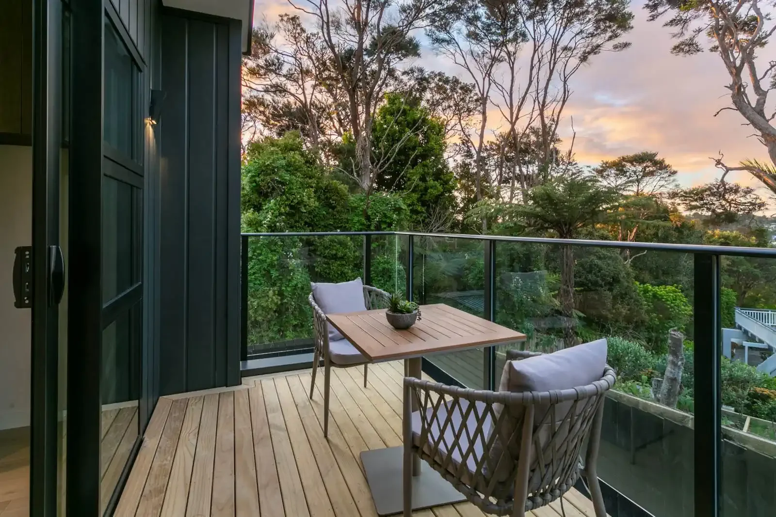 A modern outdoor balcony featuring a wooden deck, glass railings, and a small dining set overlooking lush green trees at sunset.