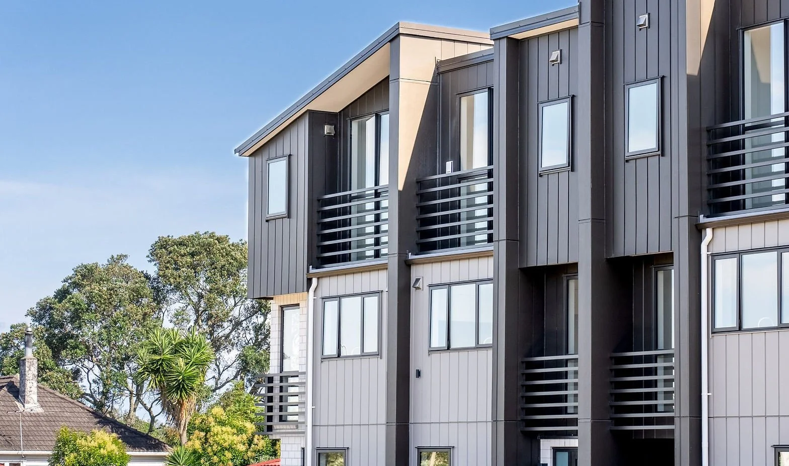 Architectural detail of a modern townhouse exterior, featuring vertical grey siding and black balcony railings against a blue sky.