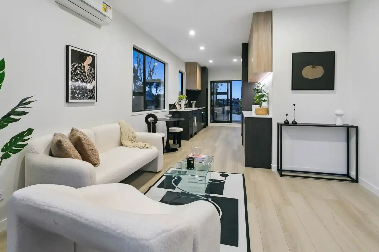 A modern open-concept living area featuring curved white sofas, light wood flooring, a geometric rug, and a sleek black kitchen.