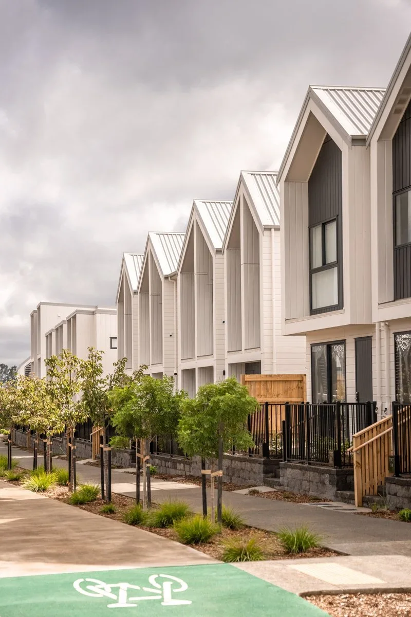 Exterior street view of a New Zealand modern new-build townhouse development featuring distinctive steeply pitched roofs, off-white vertical cladding, and landscaped front yards in Auckland.