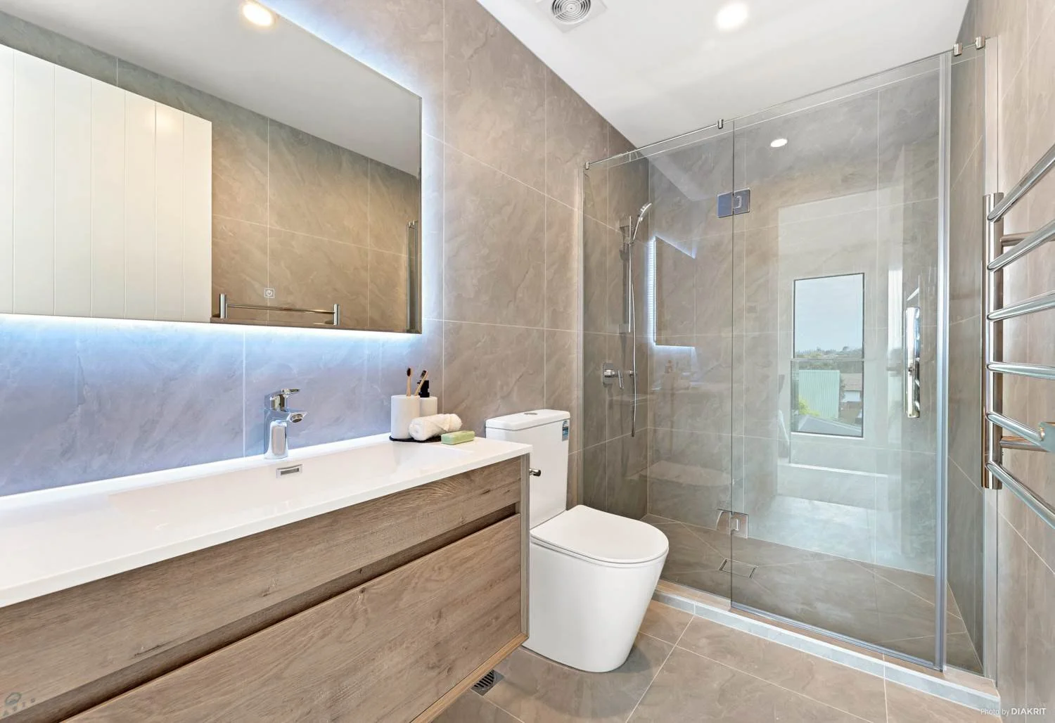 Modern bathroom interior featuring a light wood floating vanity and a frameless glass shower enclosure. The contemporary design is finished with large, elegant grey stone-look tiles on the walls and floor.