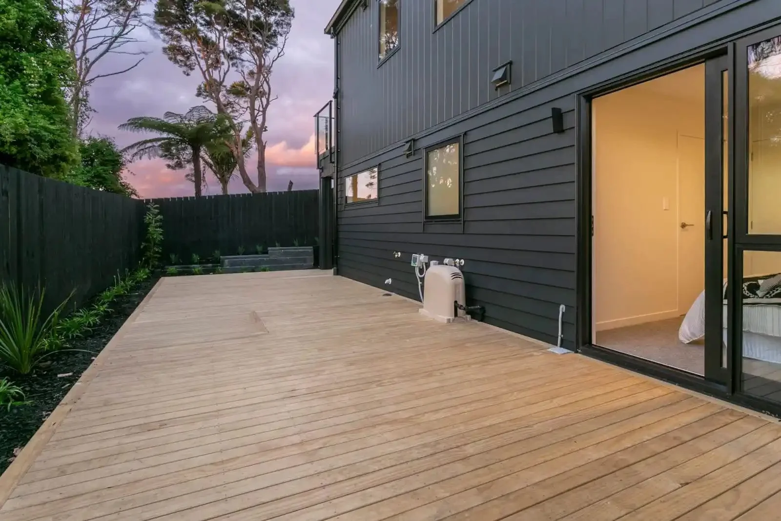 A spacious outdoor wooden deck attached to a modern dark grey house, featuring sliding glass door that open into an illuminated bedroom at dusk.