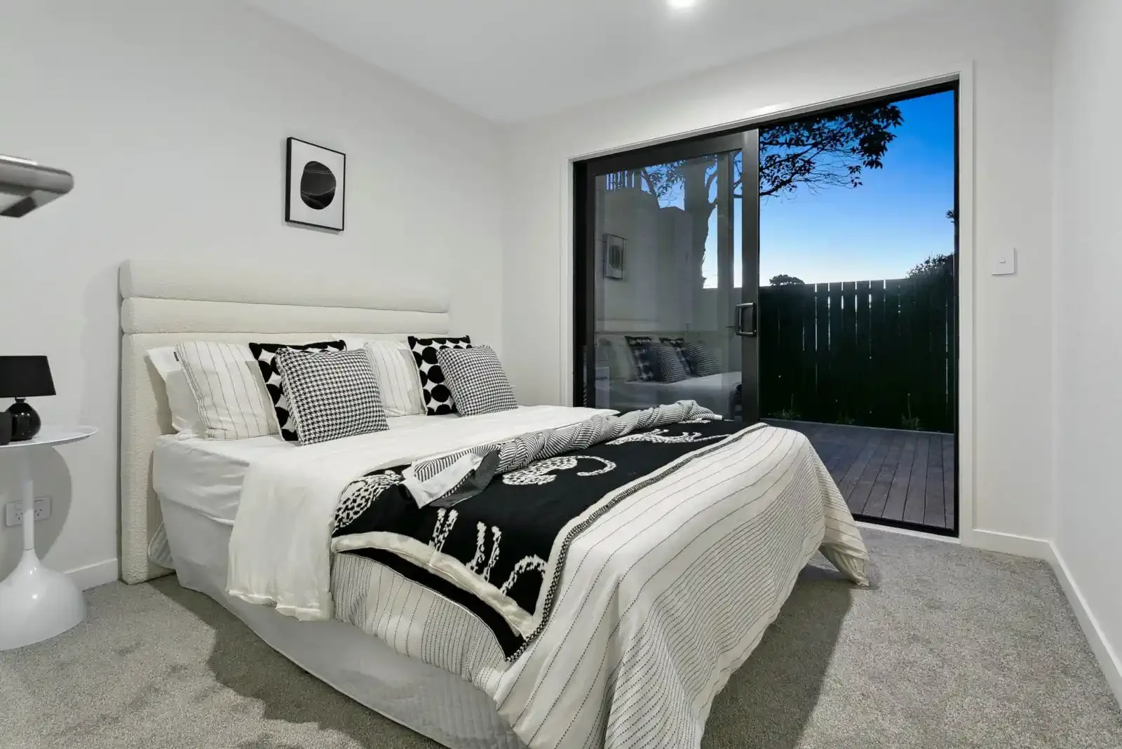 A modern bedroom featuring white walls, a bed with black and white patterned bedding, and large sliding glass doors opening onto an outdoor wooden deck.