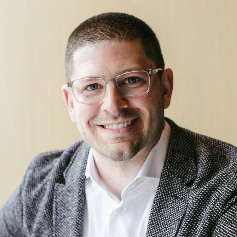 A man with glasses, short dark hair, wearing a gray patterned blazer and white shirt, smiling at the camera.
