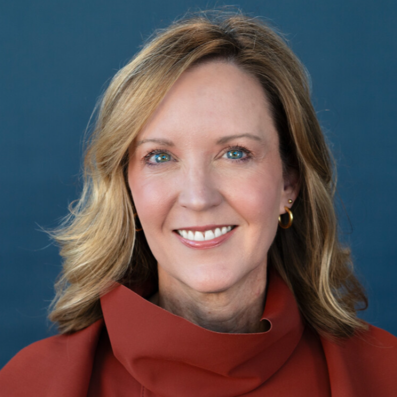 A woman with shoulder-length blonde hair, blue eyes, and a bright smile, wearing a maroon top and gold earrings, against a dark blue background.