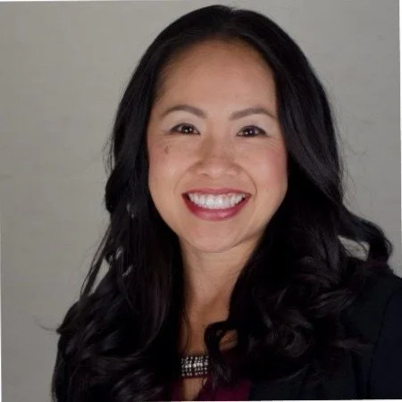 A smiling Asian woman with long black hair, wearing a black blazer and a necklace, in front of a plain light-colored background.