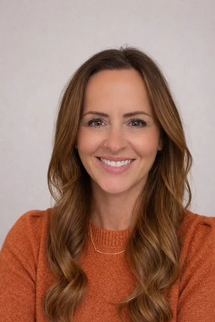 A woman with long, wavy brown hair smiling at the camera, wearing an orange sweater.