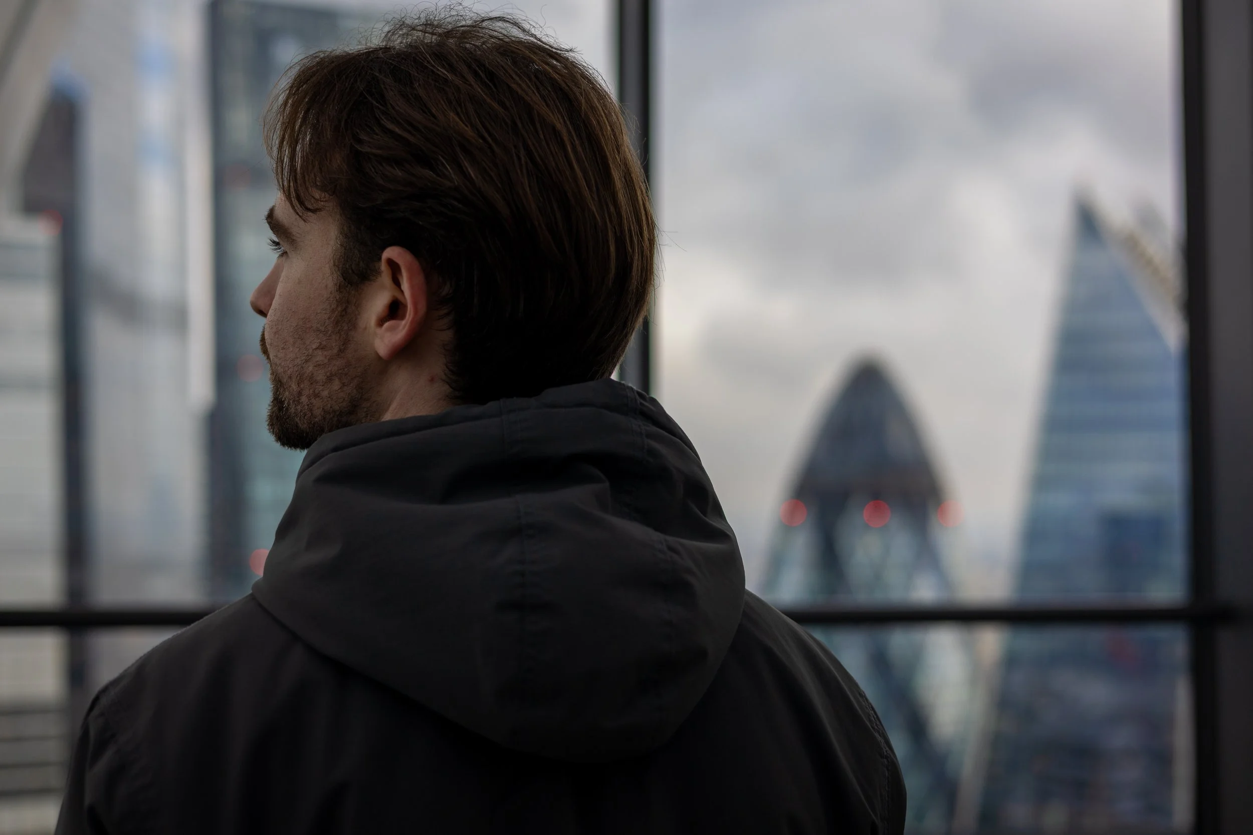 A man with brown hair and a beard wearing a black hoodie, looking out of a large window at the London skyscrapers, including the Walkie Talkie building and the Gherkin.