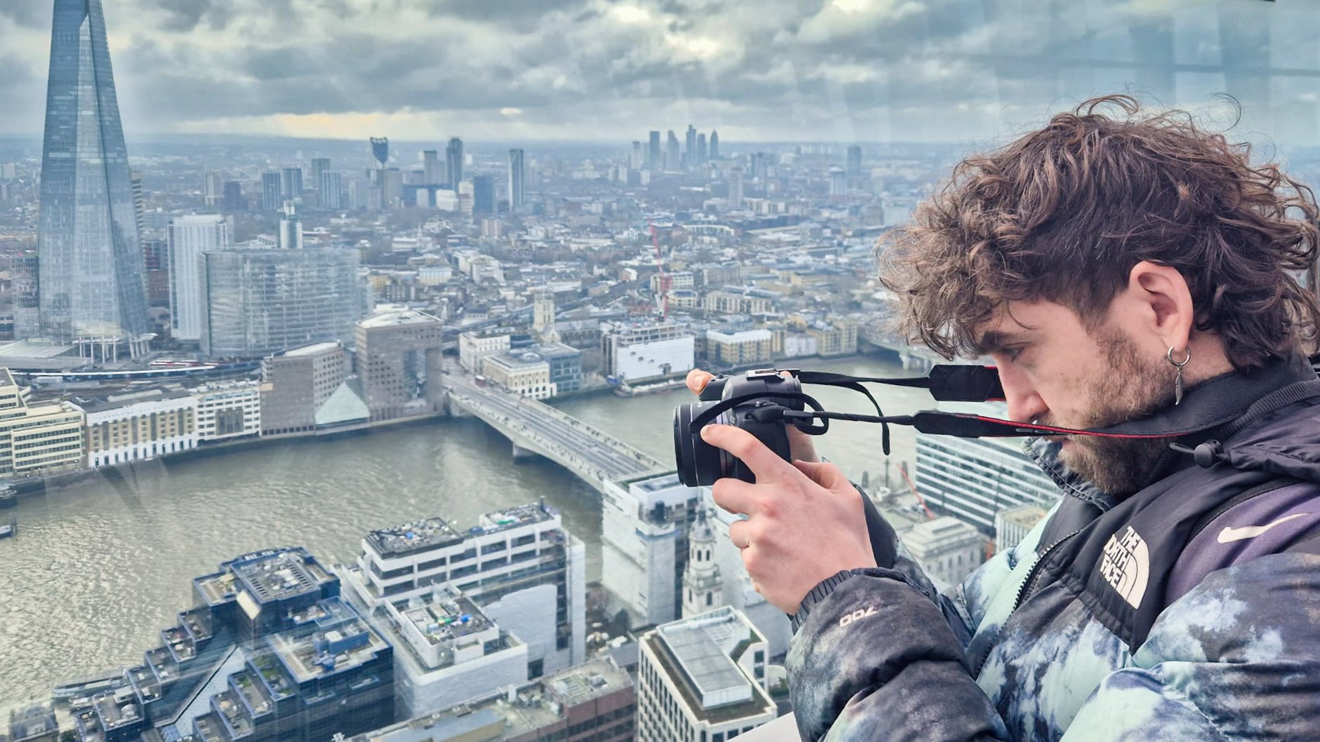 A man with curly hair and a earring is taking a photo with a camera on a high rooftop overlooking a cityscape, including the river Thames and London landmarks.