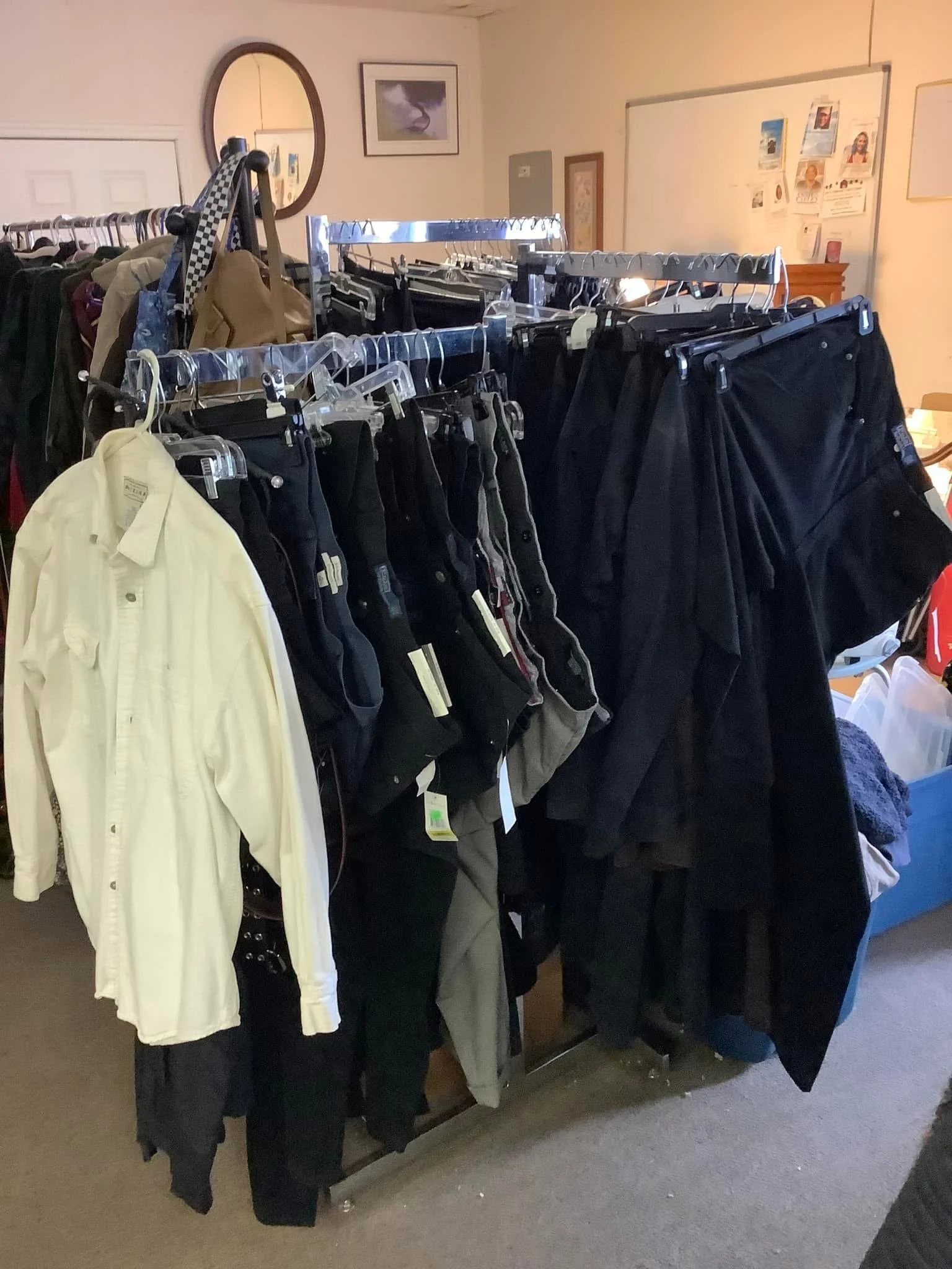 Clothing rack displaying various pants, including black, gray, and khaki, with a yellow button-up shirt hanging on the side, situated in a room with framed pictures and a mirror on the wall.