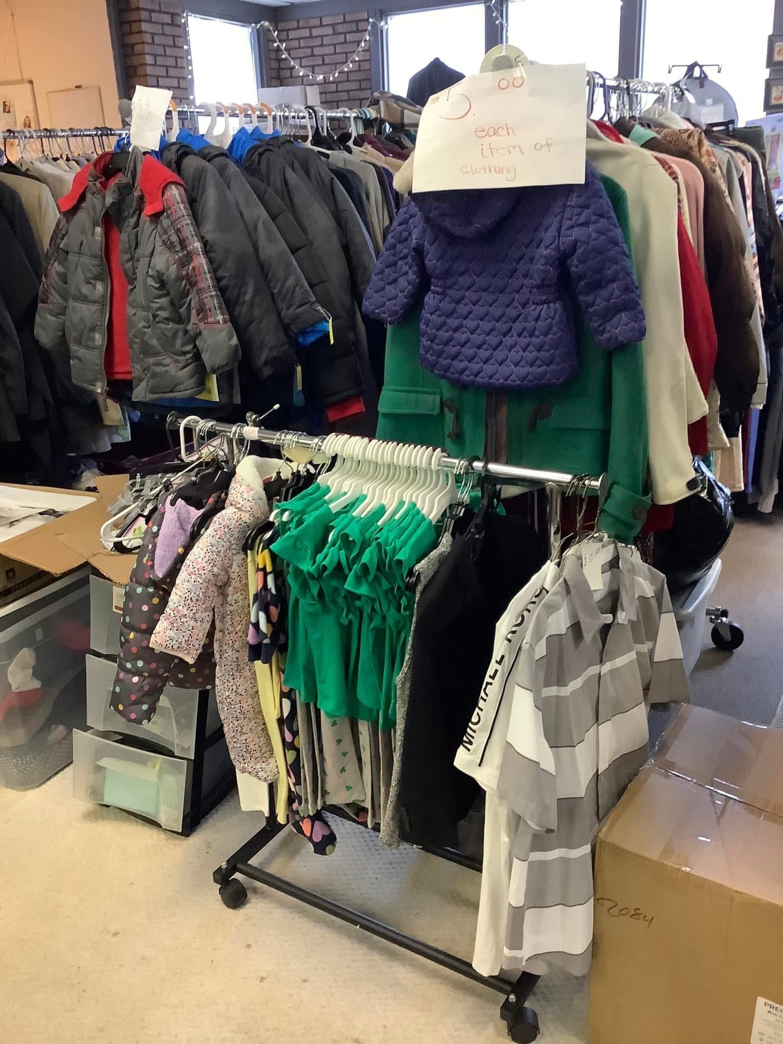 A display of children's clothing at a thrift store, including jackets, shirts, and pants on racks. A handwritten sign states, 'each item of clothing'.