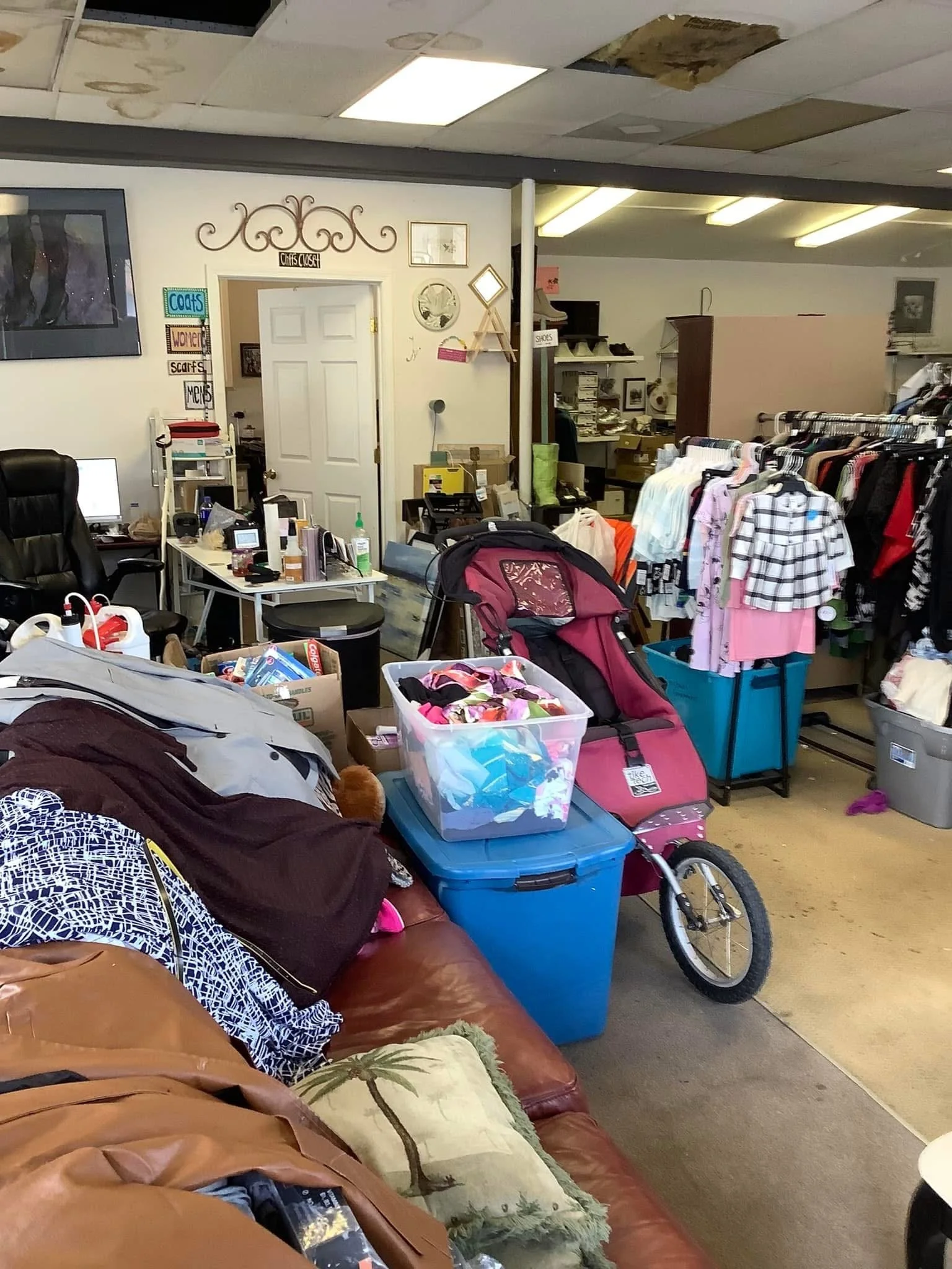 An indoor cluttered space, possibly a thrift store or donation center, with a brown leather couch and various items including a child's pink stroller, plastic storage bins, clothing racks, and miscellaneous objects.