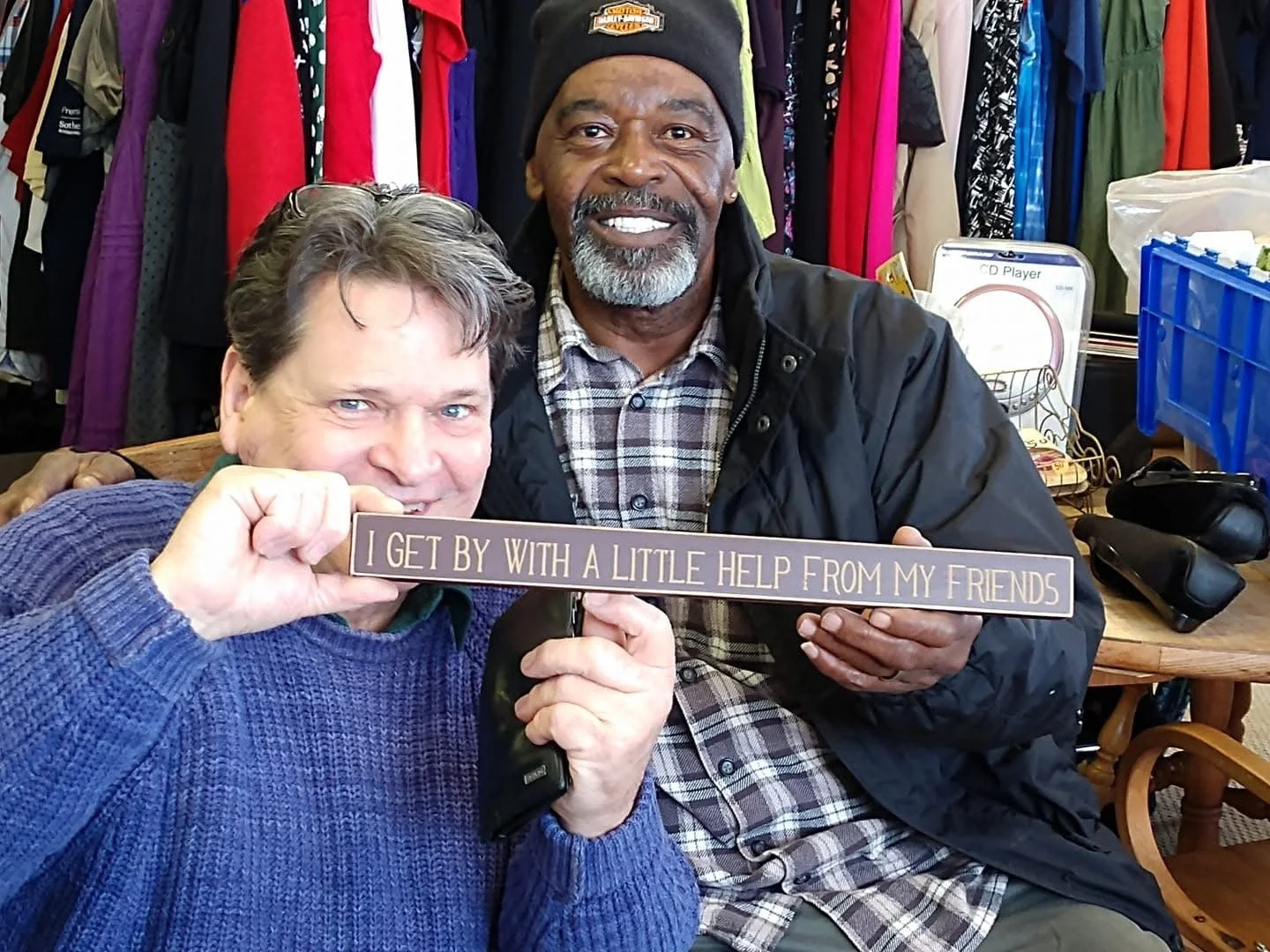 Two men smiling, sitting at a table with shelves of clothing in the background, holding a sign that reads 'I GET BY WITH A LITTLE HELP FROM MY FRIENDS'.