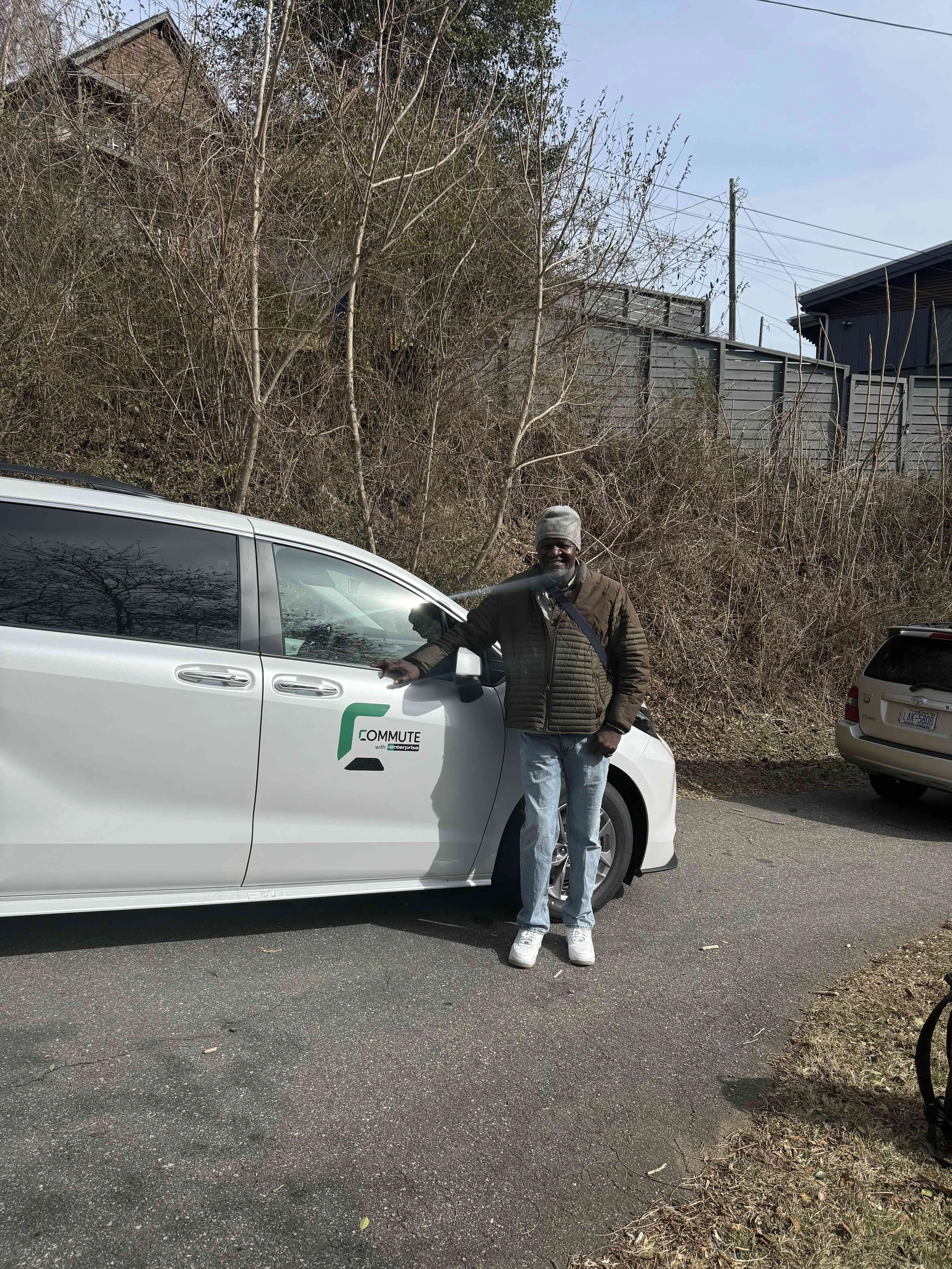 A man with gray hair, wearing a beanie, dark jacket, and jeans, standing next to a white vehicle with a 'Commute' logo on the side. The vehicle is parked on an asphalt surface, with a background of leafless trees, bushes, and utility poles.