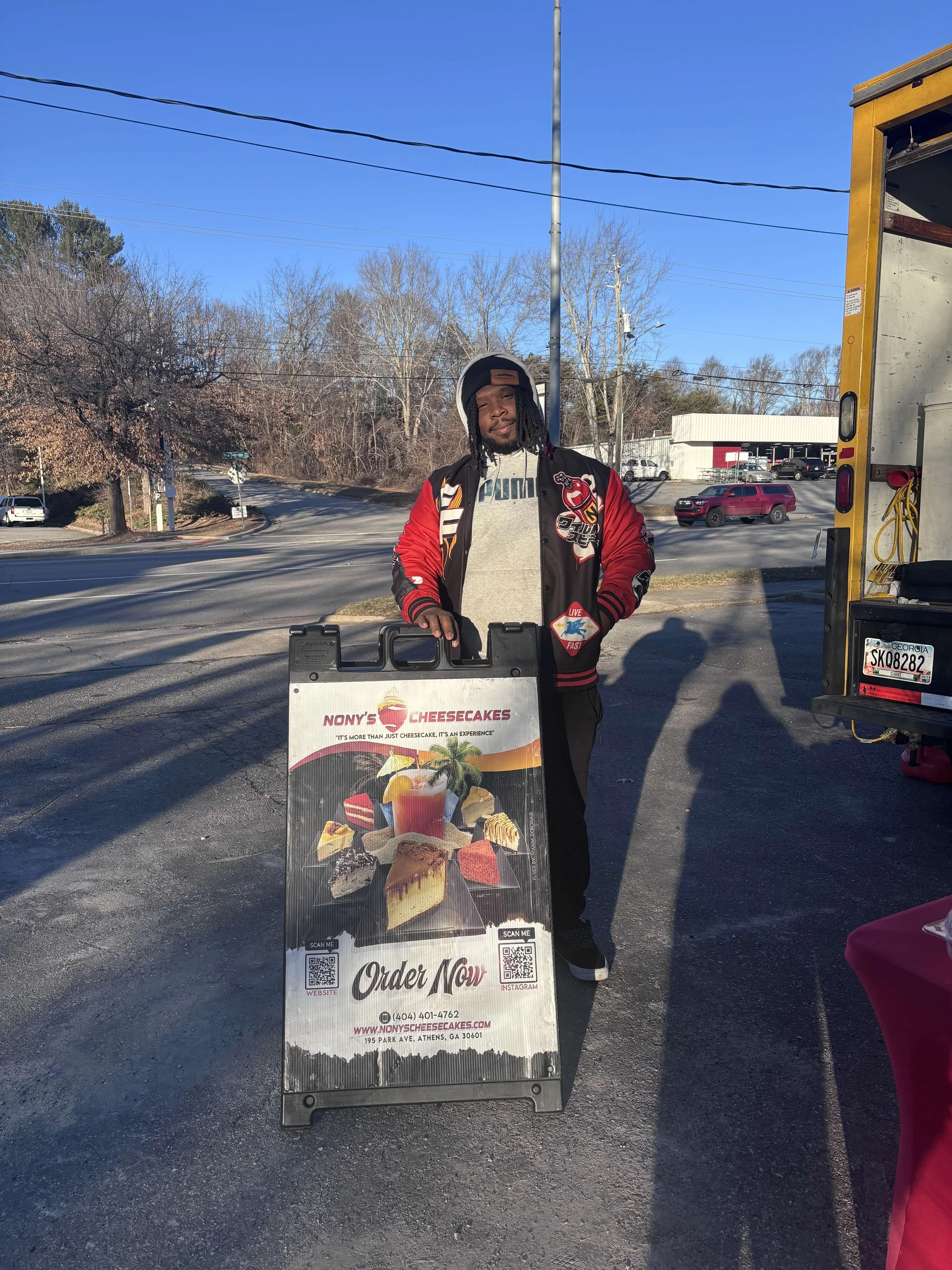 Young man standing outside holding a signboard for Nony's Cheesecakes, with a food truck nearby and a street with cars in the background on a sunny day.
