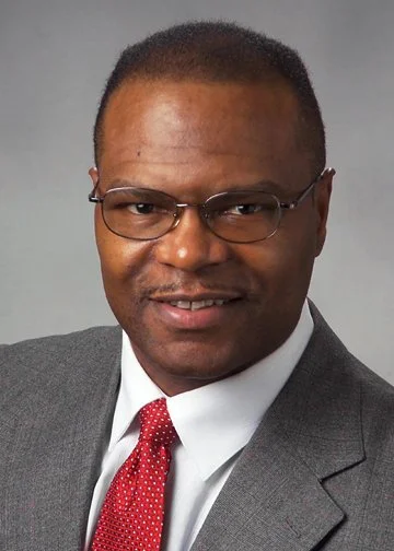 A man in a gray suit, white shirt, and red tie, wearing glasses, smiling at the camera.