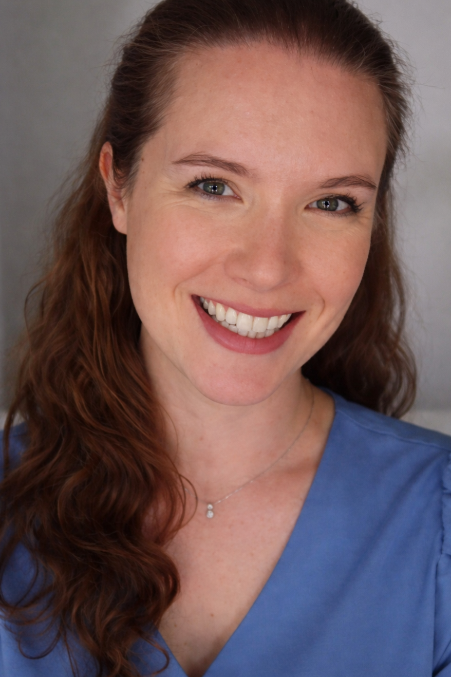 A smiling woman with long wavy red hair, green eyes, and light skin, wearing a blue top and a delicate necklace, against a neutral background.