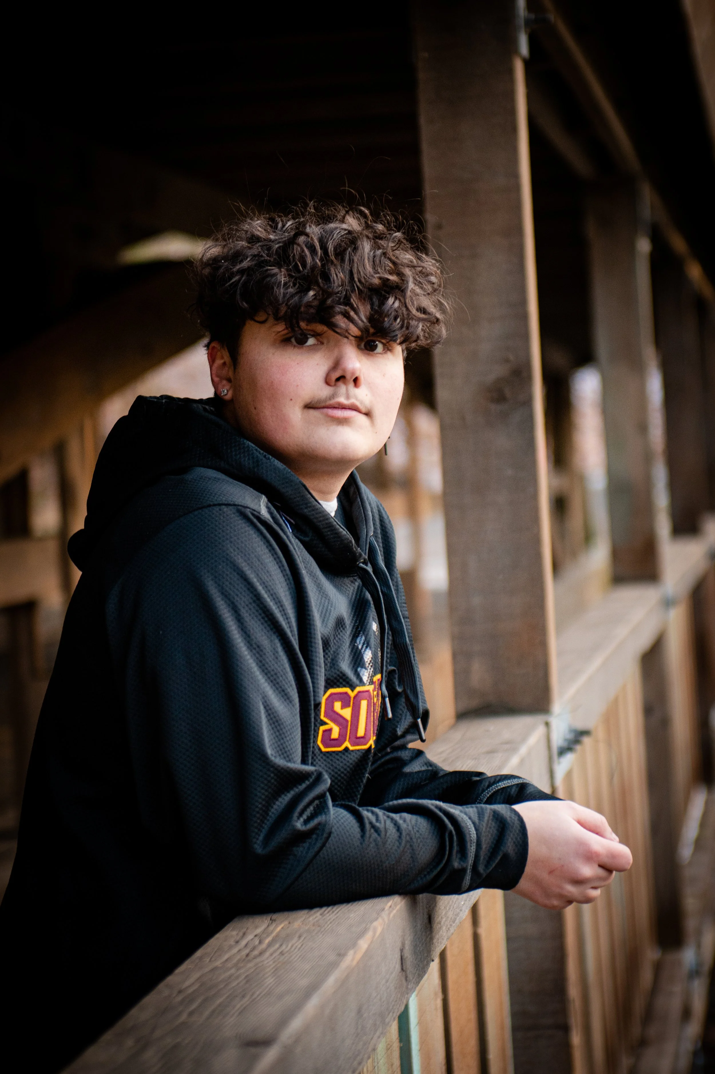 A young person with curly brown hair, wearing a black hoodie with some letters in yellow and red, is leaning on a wooden railing under a bridge or outdoor structure, looking at the camera.