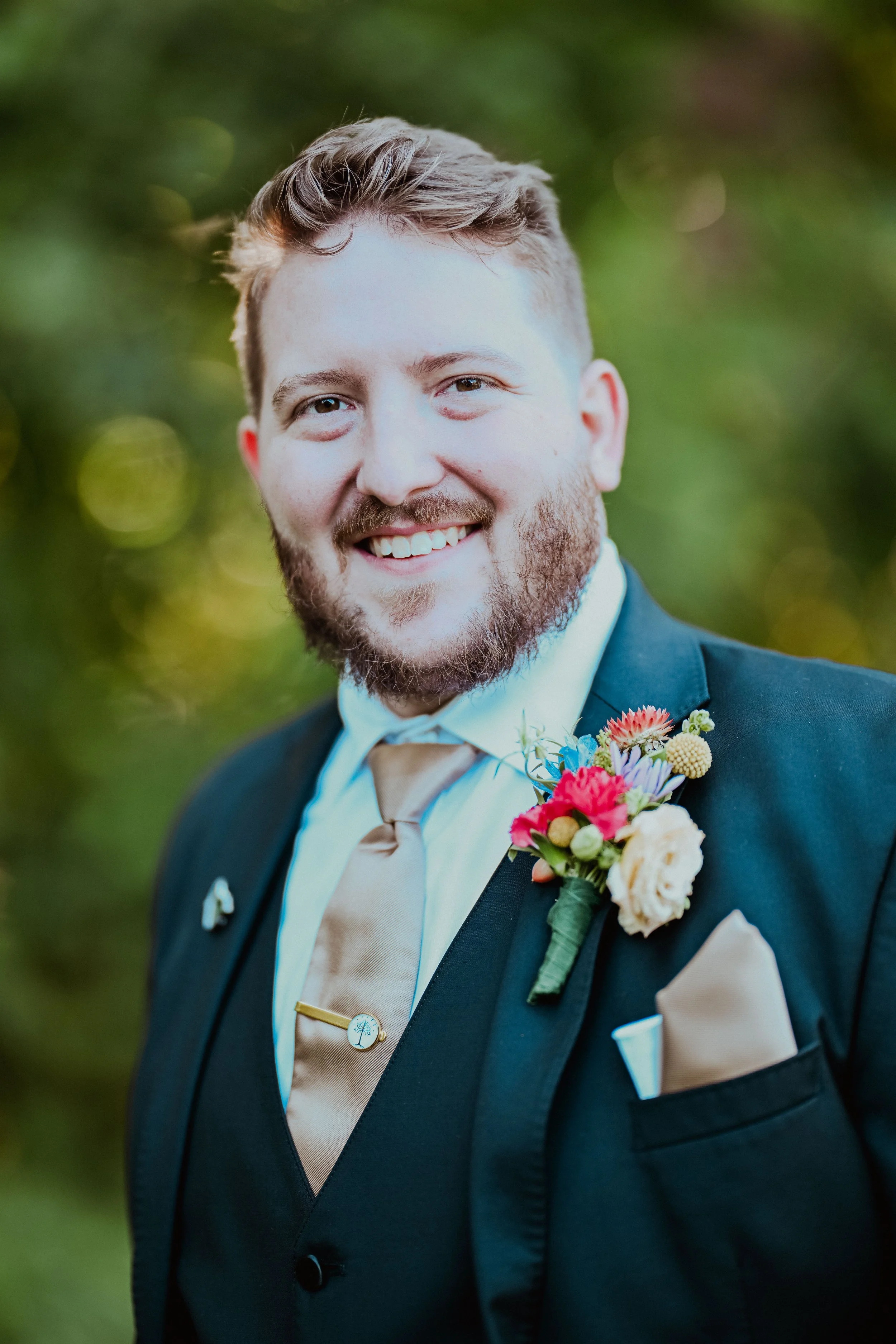 A happy man dressed in a dark suit with a beige tie and a cream-colored pocket square, wearing a boutonniere with colorful flowers, smiling outdoors with blurred green foliage in the background.