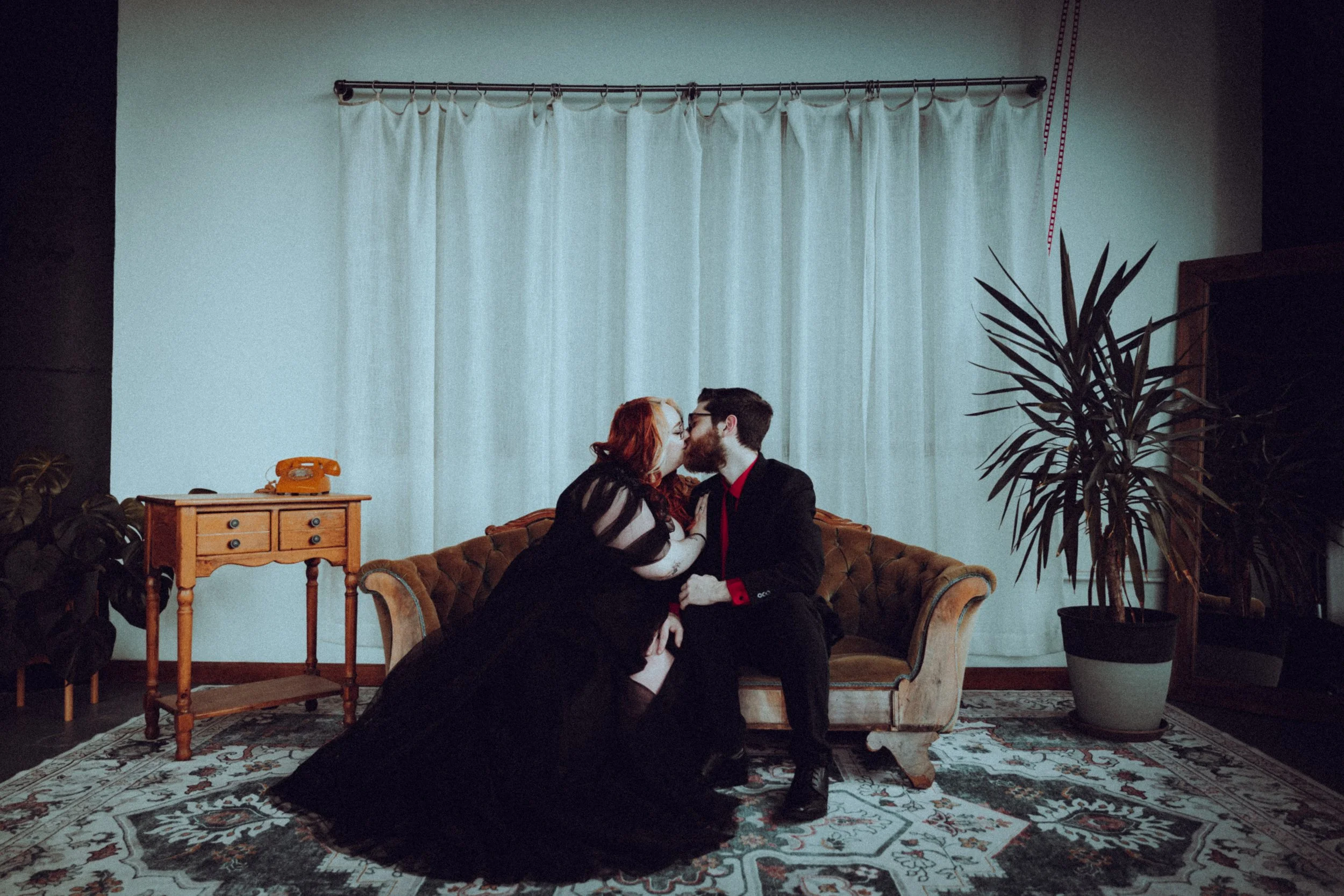 A couple sitting on a vintage sofa, sharing a kiss in a living room with a large potted plant, a wooden side table with an orange rotary phone, and white curtains.