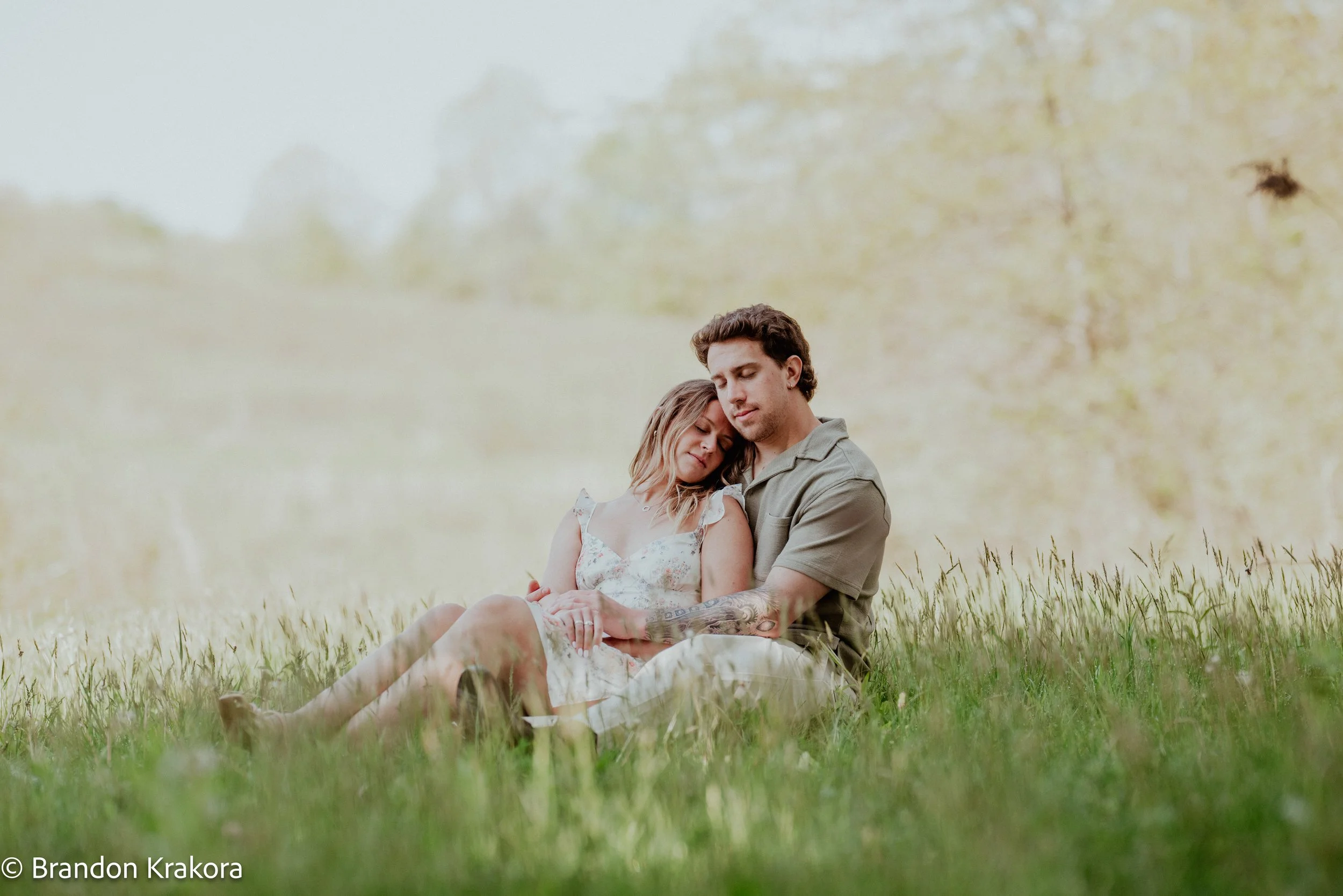 A young couple sitting on a grassy field, with the woman resting her head on the man's shoulder as they both look down.