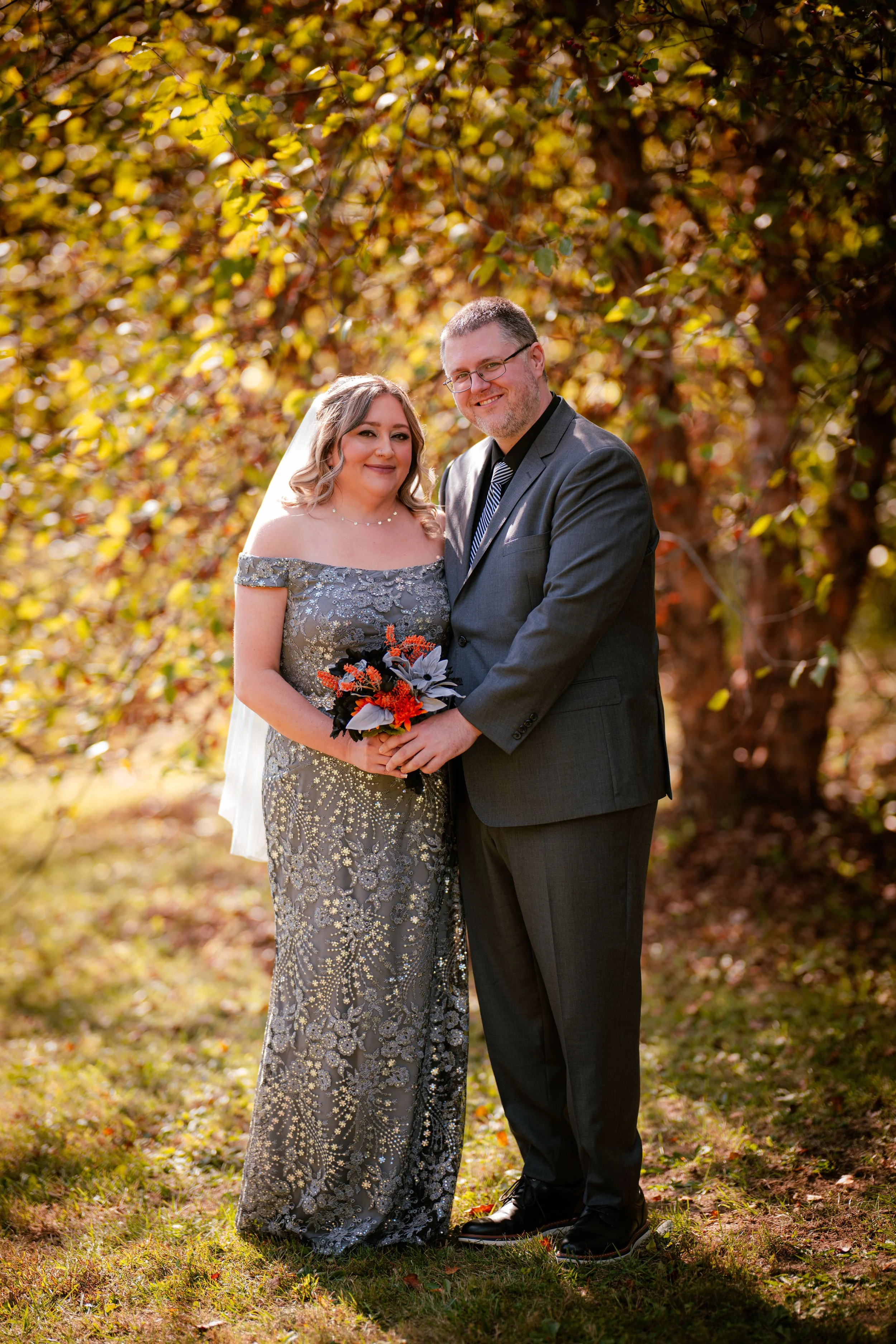 A bride and groom standing outdoors during autumn, holding hands and smiling at the camera, with trees and fall foliage in the background.
