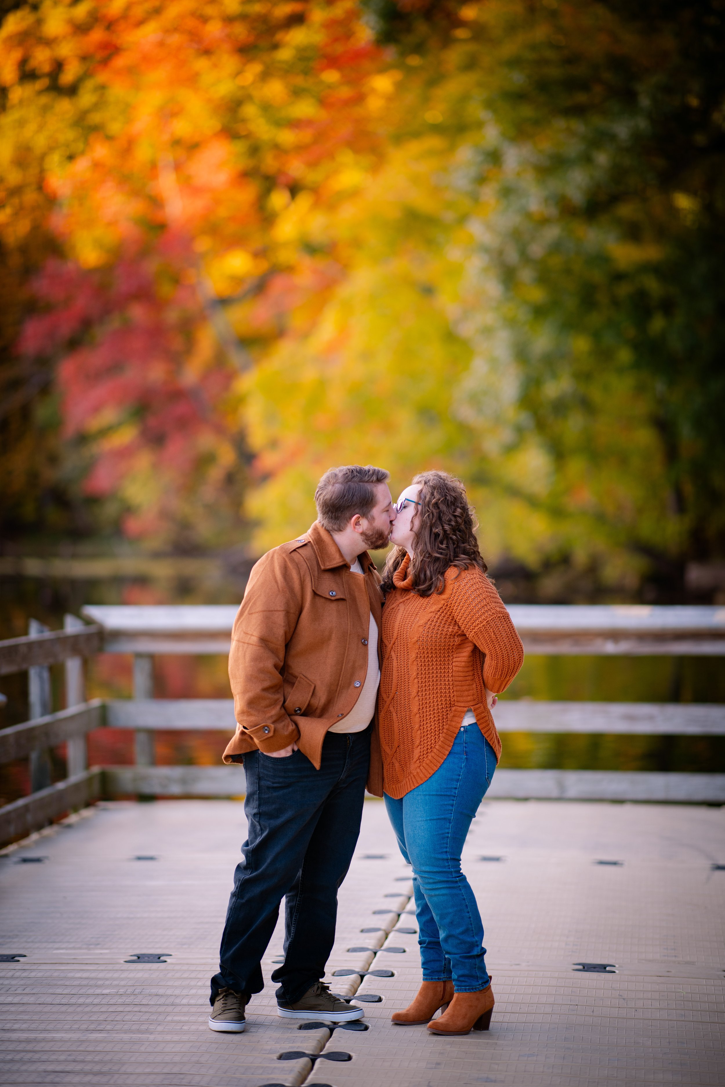 A couple kissing on a wooden dock with colorful autumn trees in the background.