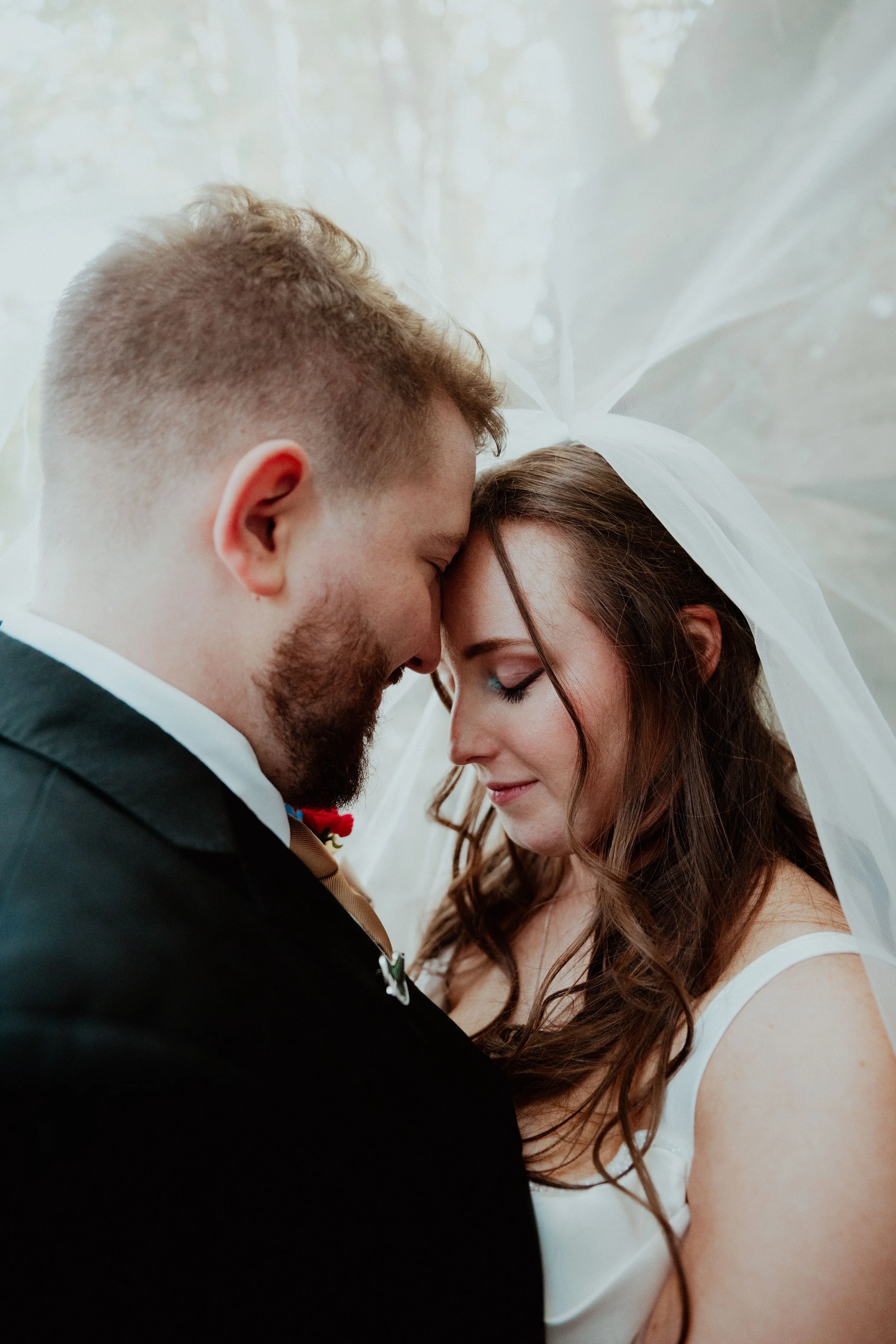 A bride and groom close together, their foreheads touching, under a white veil, with eyes closed and gentle smiles.
