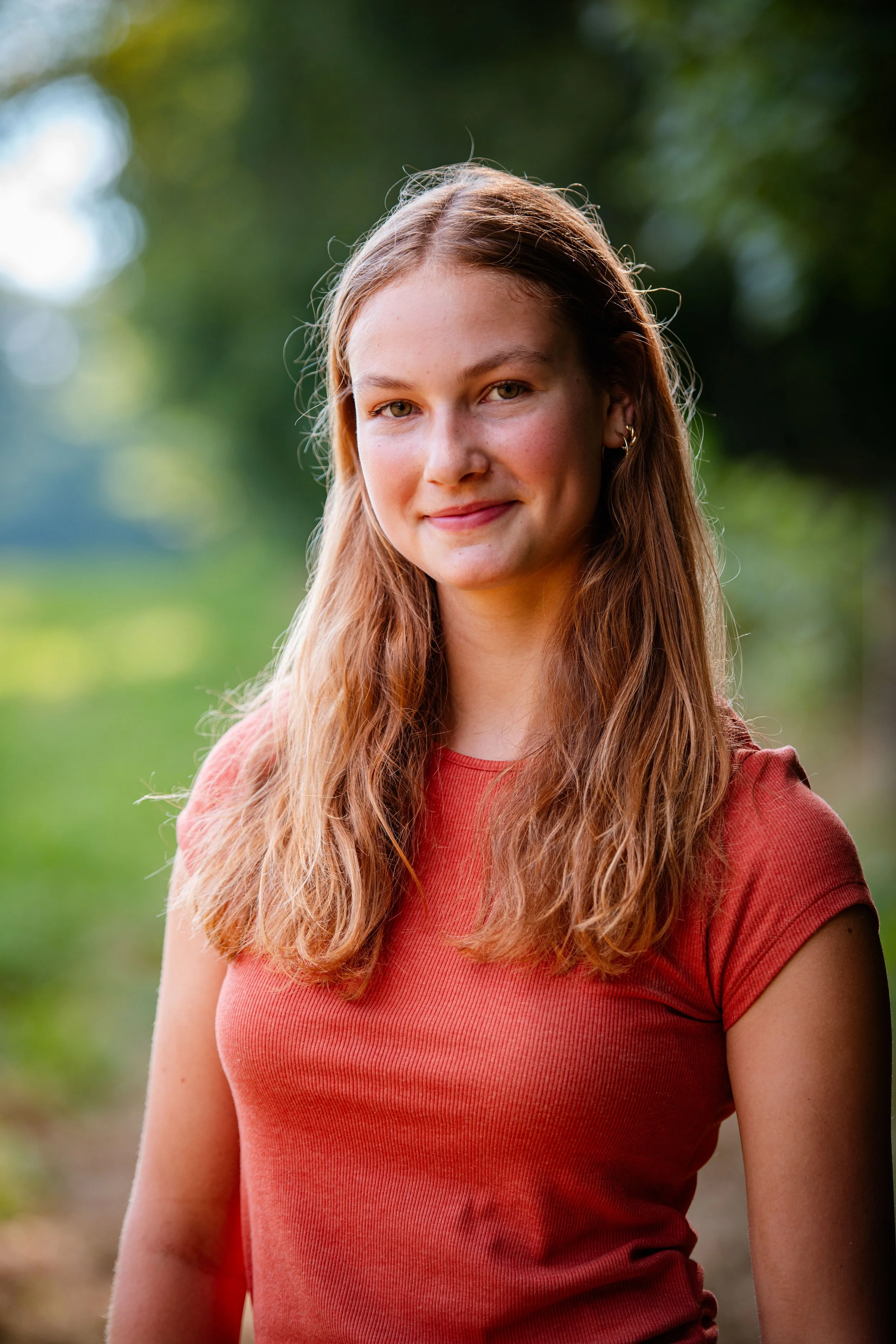 Young woman with long wavy red hair wearing a coral t-shirt outdoors with a blurred green background.